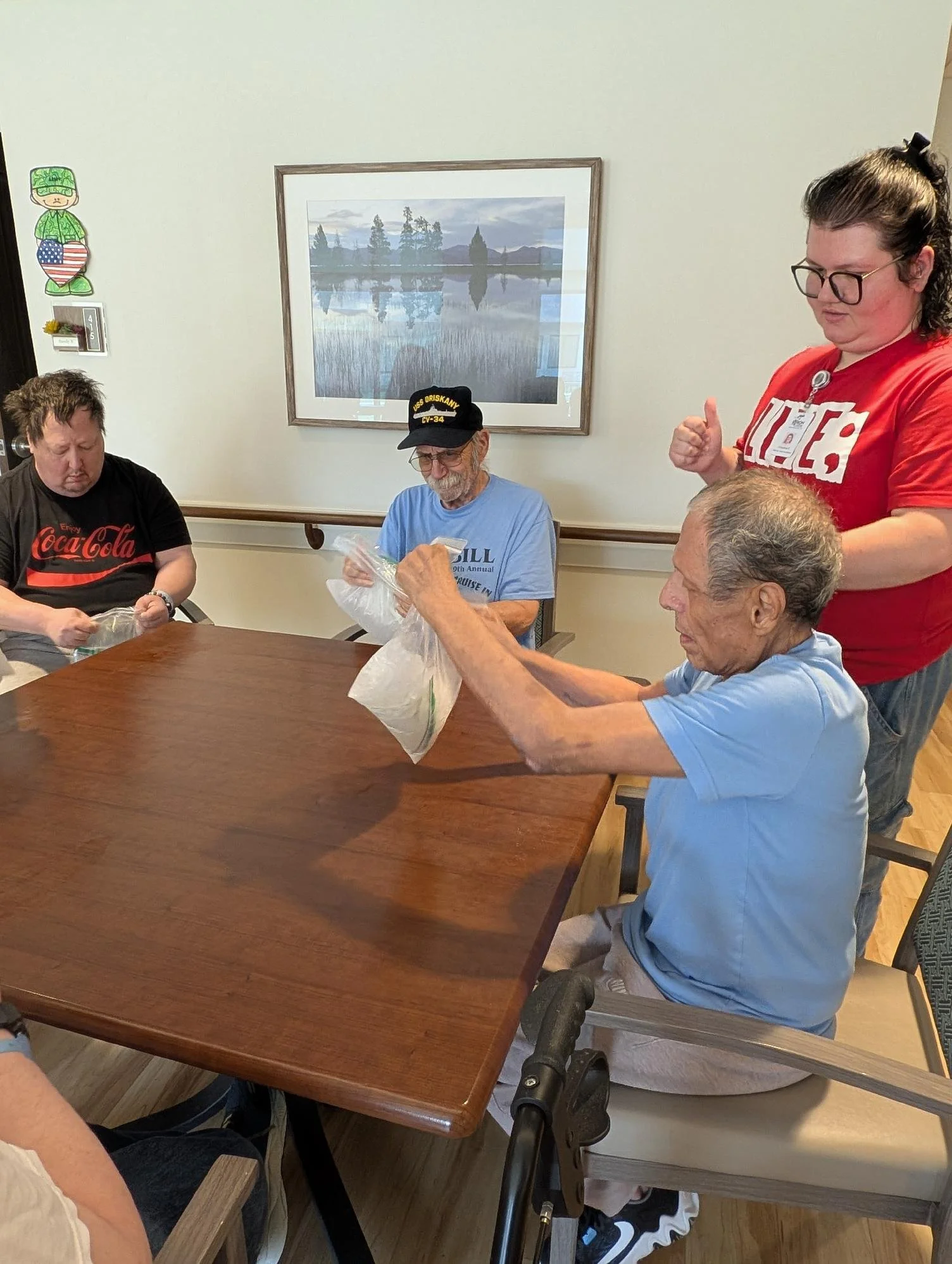 A group of elderly men seated around a wooden table, with one man in a wheelchair, and a woman standing beside them in a dining or activity room. The room has a large framed landscape picture on the wall and various decorations.