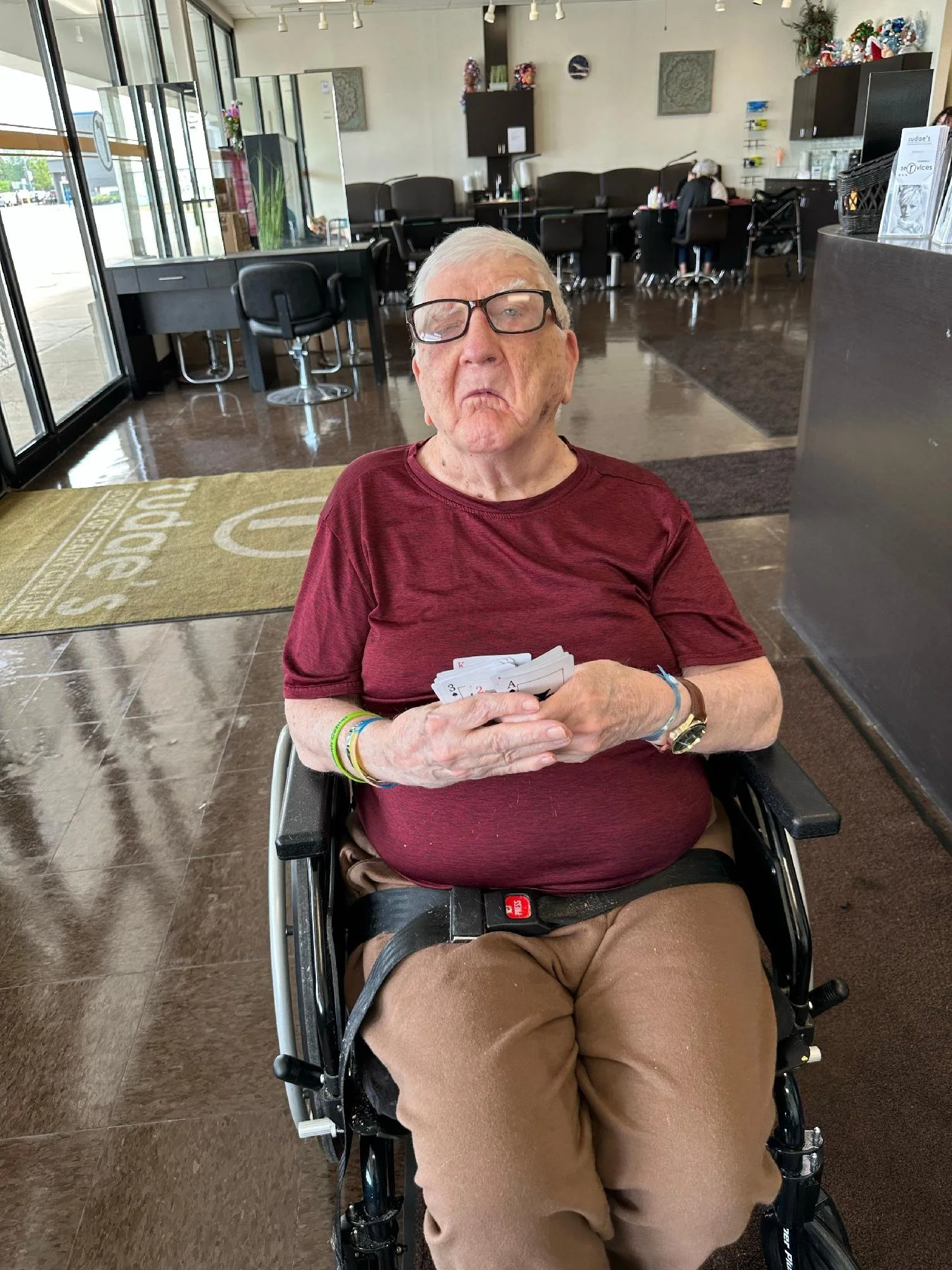 An elderly man sitting in a wheelchair inside a restaurant or cafe, holding playing cards in his hand, with a neutral or slightly disgruntled facial expression, wearing glasses, a maroon t-shirt, and brown pants.