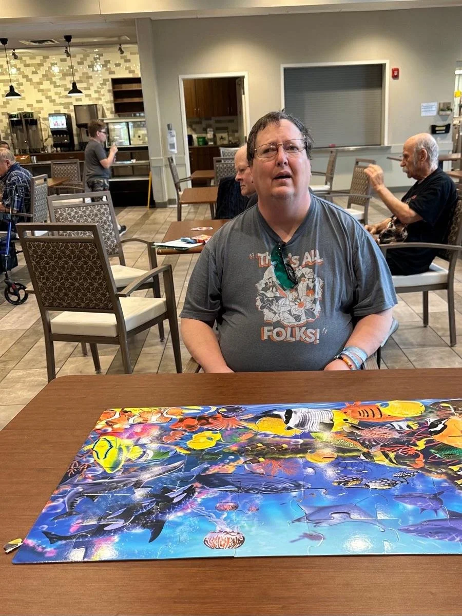 A man sitting at a table with a colorful underwater-themed jigsaw puzzle in front of him in a cafe or restaurant setting. Other people are seated at tables in the background, and a server is behind the counter.
