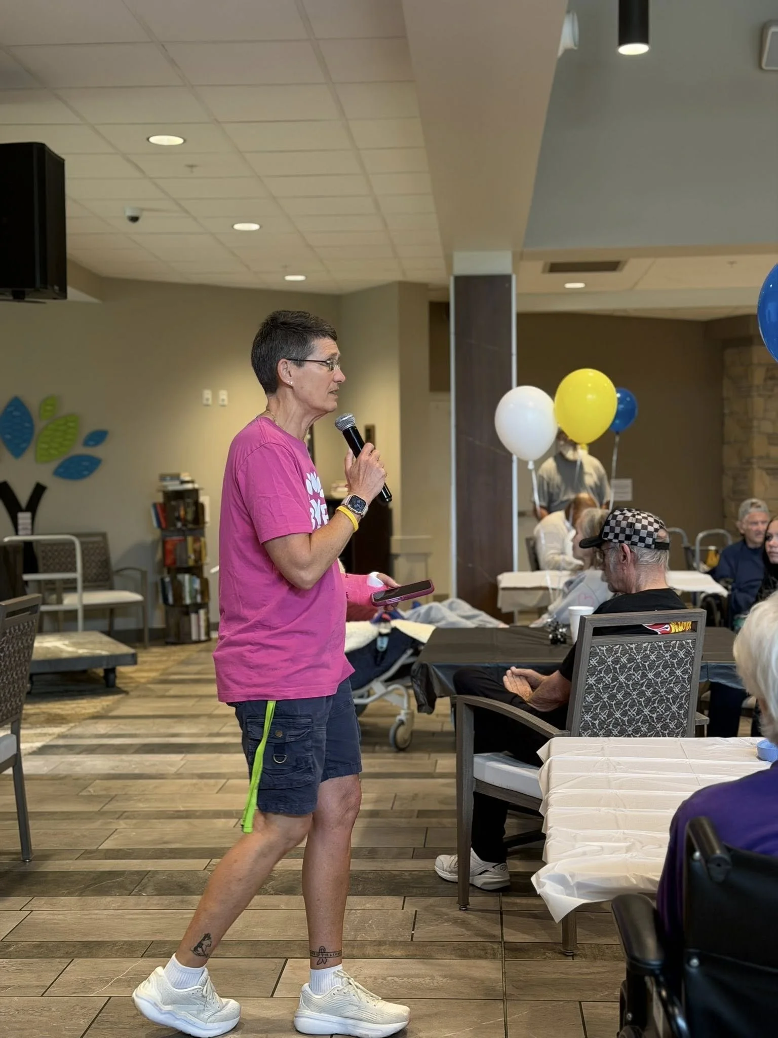 A person with short dark hair, glasses, and tattoos on their legs, wearing a pink shirt, dark shorts, and big white sneakers, holding a microphone and a phone, standing in a room with people sitting at tables decorated with balloons.