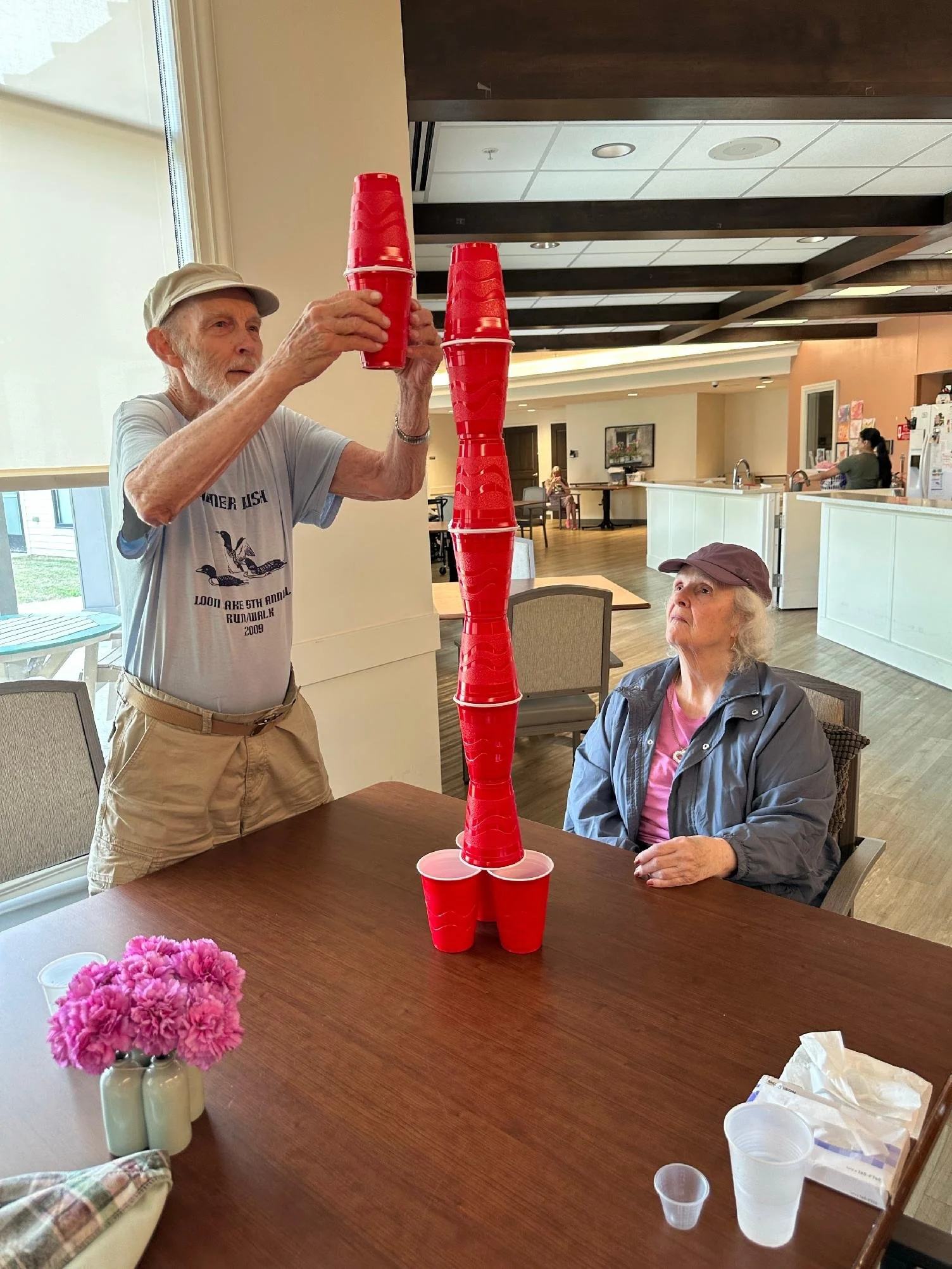 An elderly man and woman playing a stacking game with red plastic cups in a dining area. The man is stacking cups on a table while the woman watches. There is a pink potted flower on the table and a window with sunlight coming through.