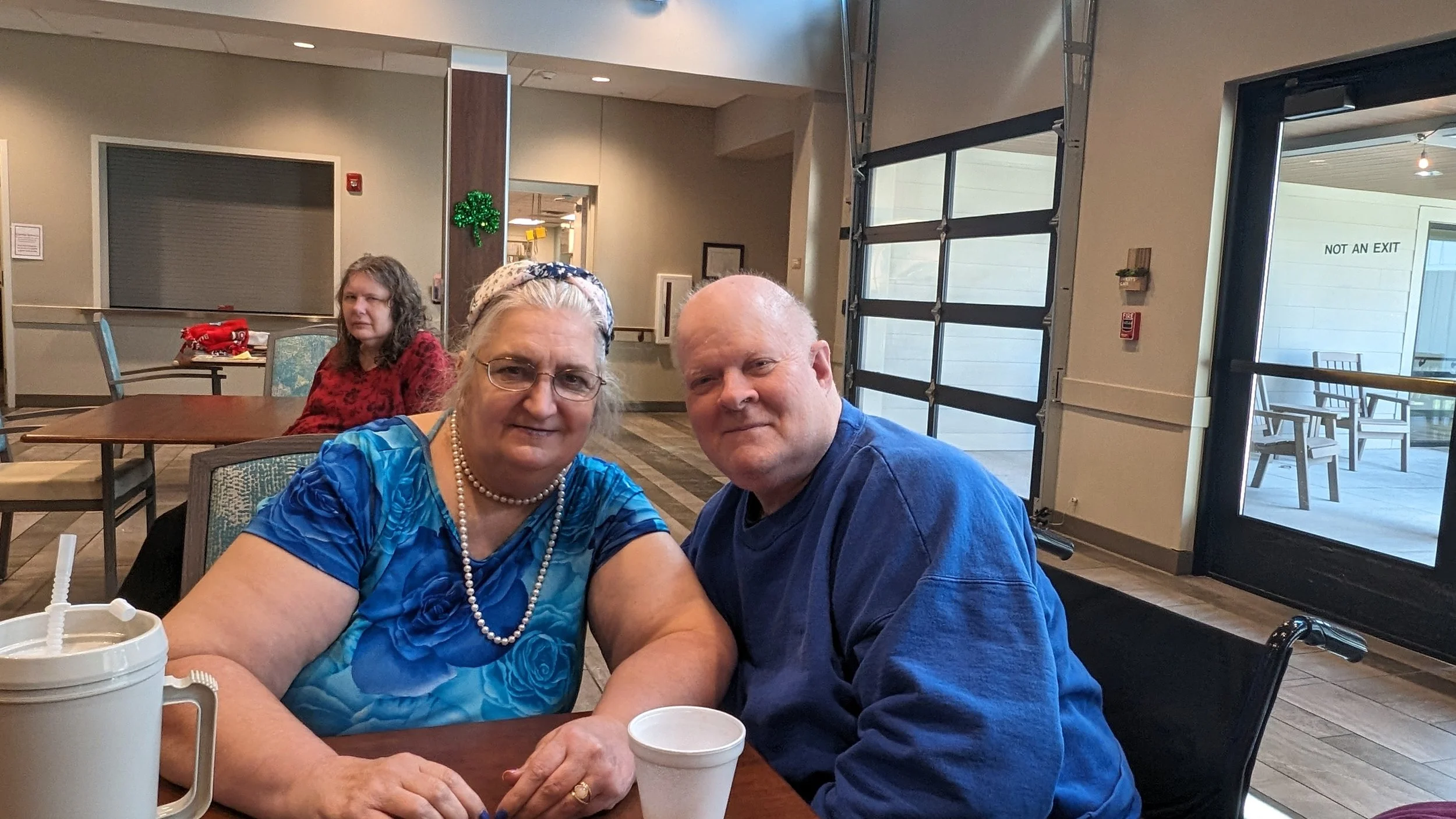 A group of three people sitting at a table in a casual restaurant or café. Two are in focus in the foreground: an older woman with glasses, a pearl necklace, and a headband, and an older man with a bald head, both smiling at the camera. A younger woman with curly hair and glasses is sitting in the background, looking slightly away. The table has drinks and a straw. The setting is indoors with large windows and a door with a sign that says 'NOT AN EXIT'.