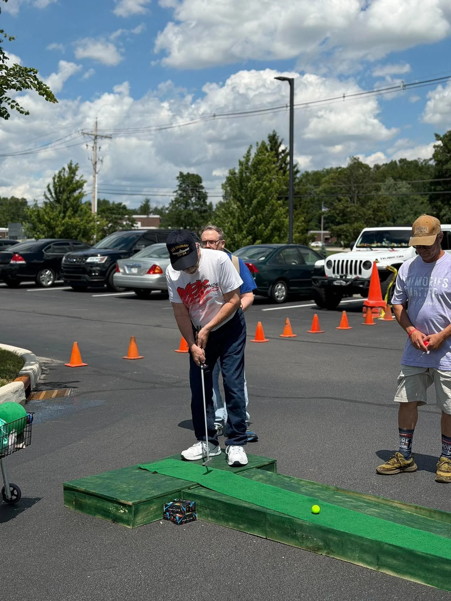 People participating in a mini-golf game outdoors in a parking lot under a partly cloudy sky. One person is lining up a shot on a green mini-golf course with orange cones and a golf ball.
