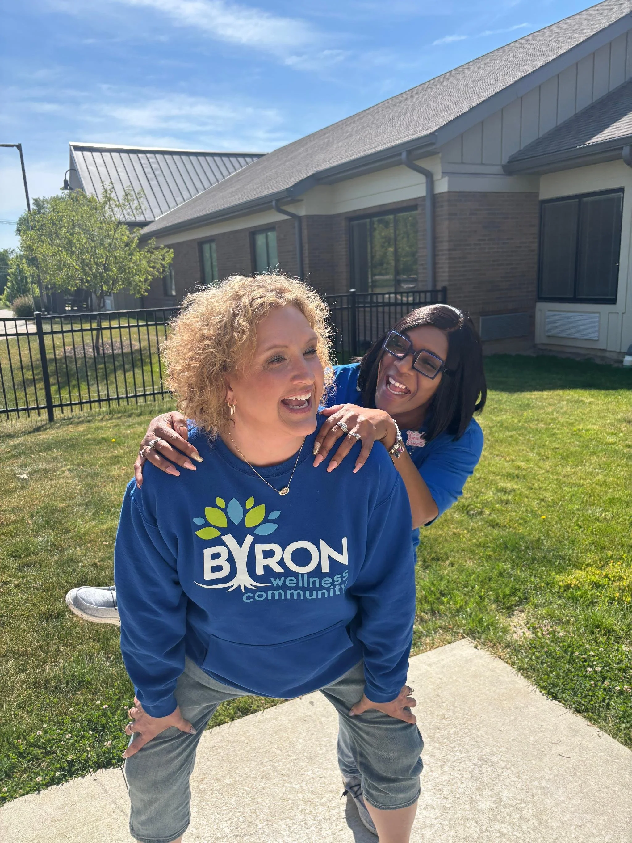 Two women smiling, one kneeling with hands on knees, the other standing behind with hands on the kneeling woman's shoulders on a sunny day in front of a house with a brick and siding exterior, a tree, and a black metal fence.