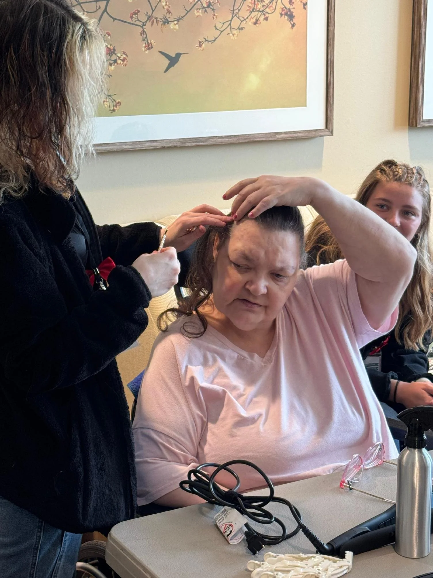 A woman with long blonde hair sitting at a table, receiving a haircut or hair treatment from another woman with long curly hair. There are three people visible, and they are in a room with paintings or framed pictures on the wall.