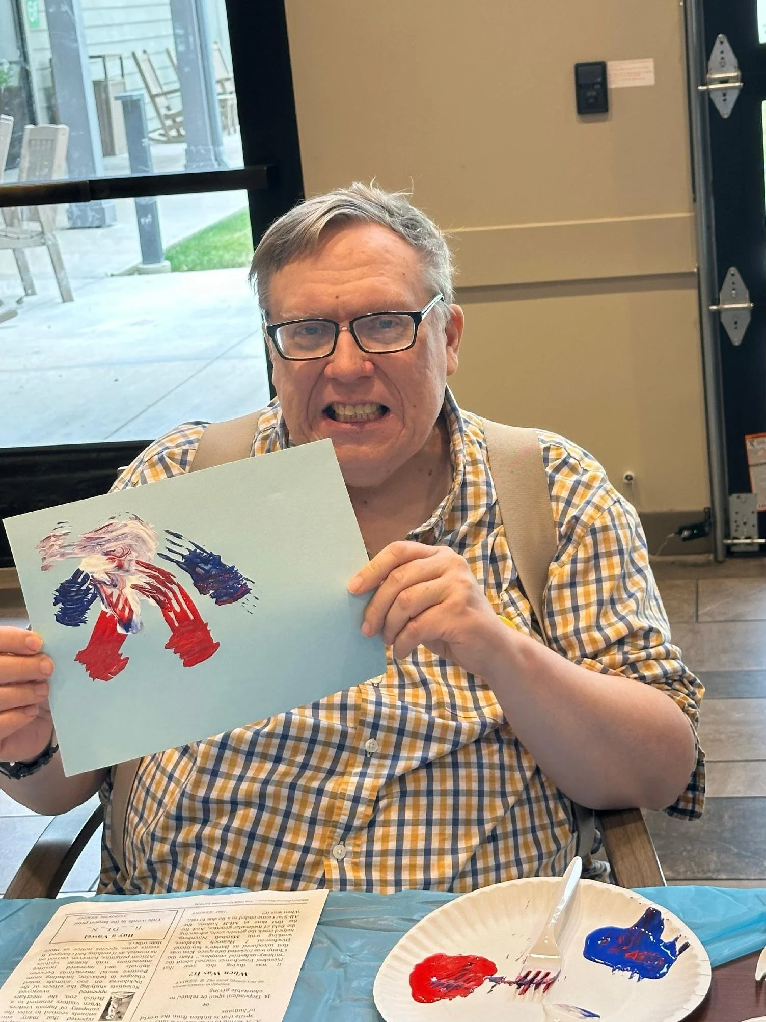 A man with glasses and a checkered shirt makes a grimace while holding a piece of paper with colorful painted figures. The table in front of him has paint on a paper plate, a fork, and a piece of paper with text.
