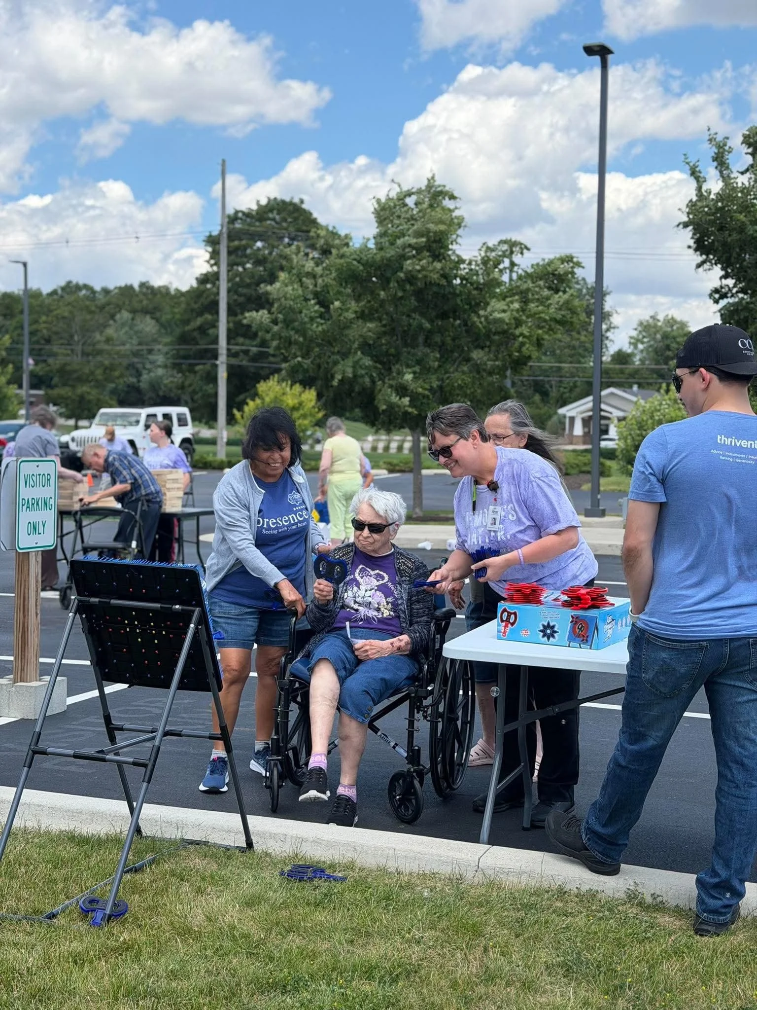 A group of people gathered outdoors in a parking lot, some are standing and others are seated, with tables and items for an event or gathering. There is a clear sky with some clouds, trees, and a few cars in the background.