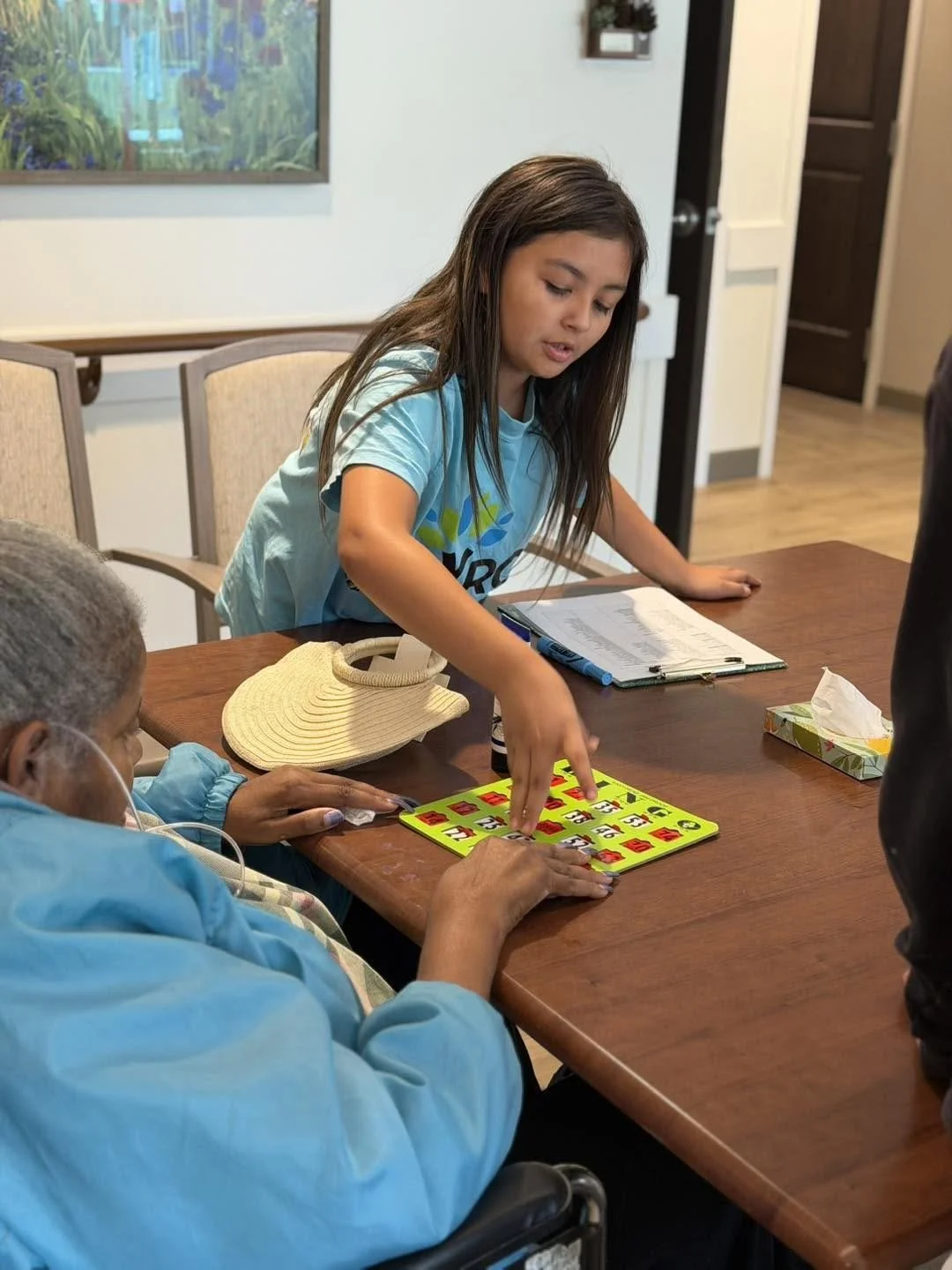 A young girl and an elderly woman playing bingo at a wooden table in a room with chairs, a painting on the wall, and a tissue box.