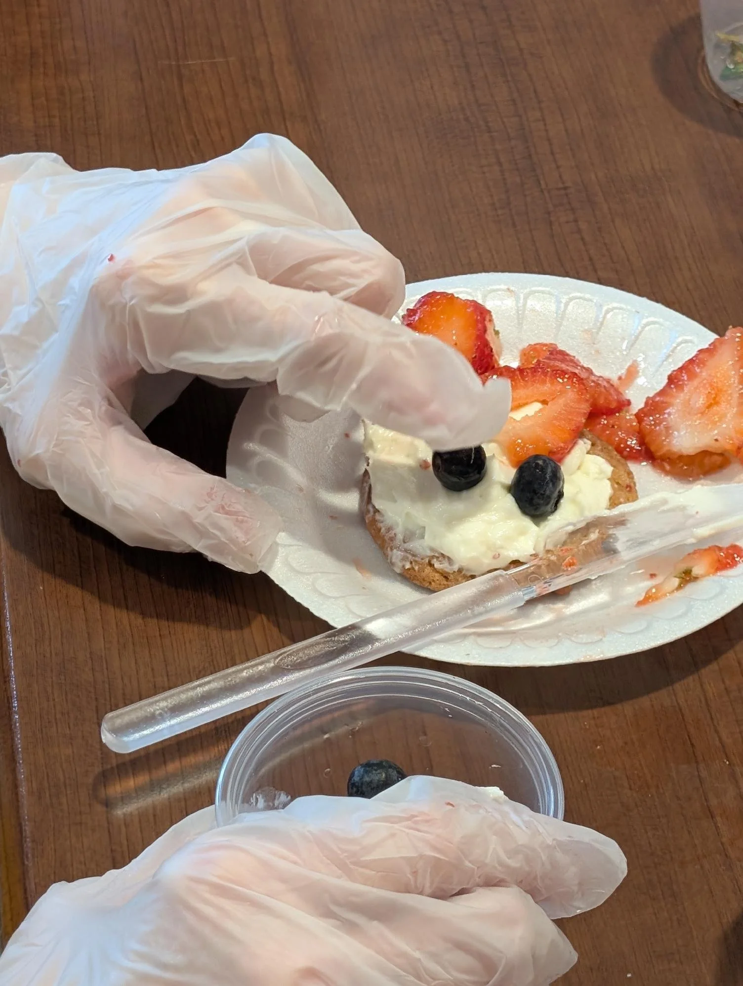 A hand wearing a disposable glove is holding a piece of cake with strawberries and blueberries on top. There are additional strawberries and blueberries on the cake, which is on a white paper plate. Part of a plastic container and a plastic utensil a