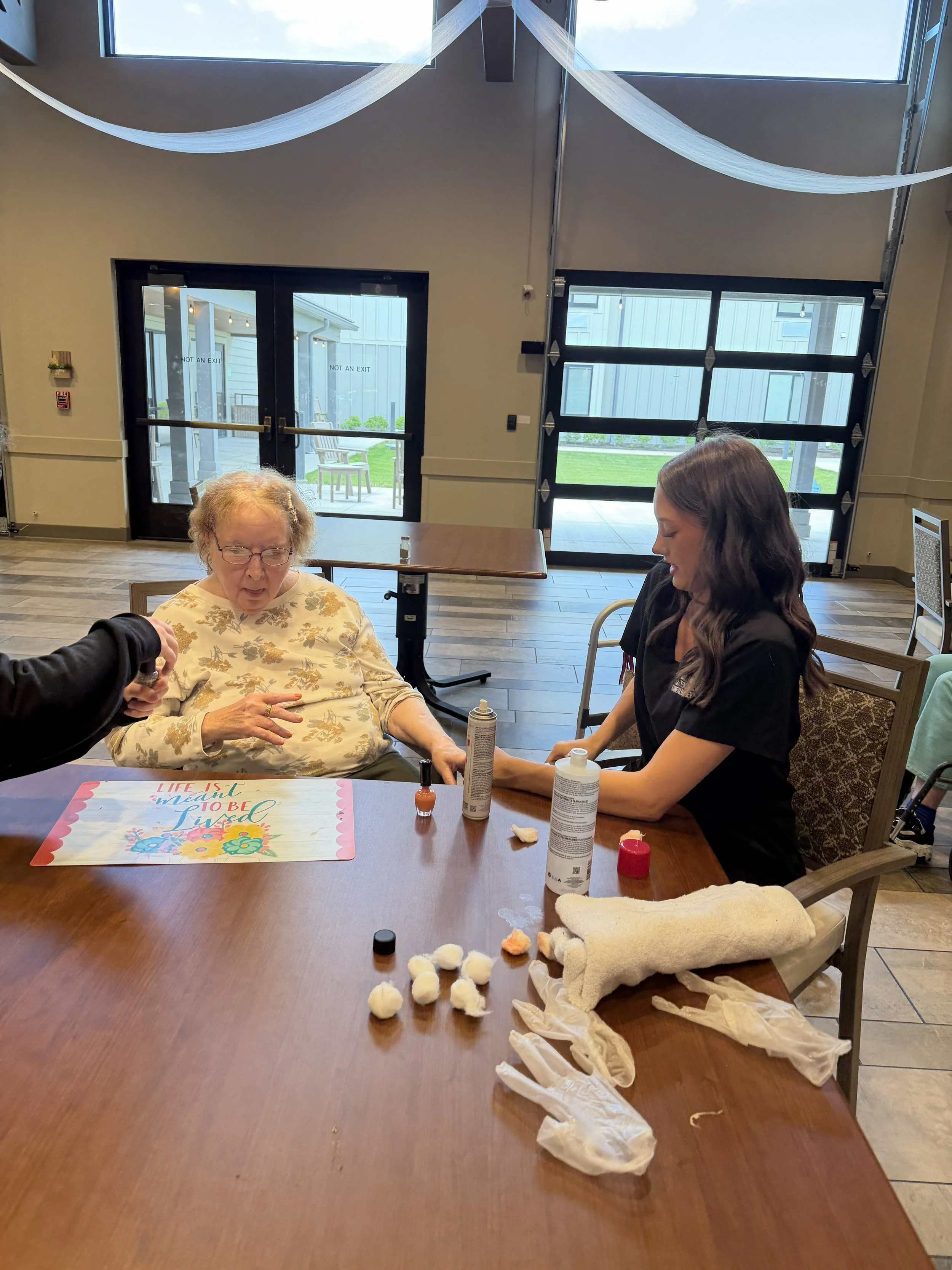 A woman in a floral shirt with glasses and an elderly woman in a black shirt are sitting at a table engaging in a craft activity, surrounded by various craft supplies including cotton balls, bottles of lotion, and gloves.