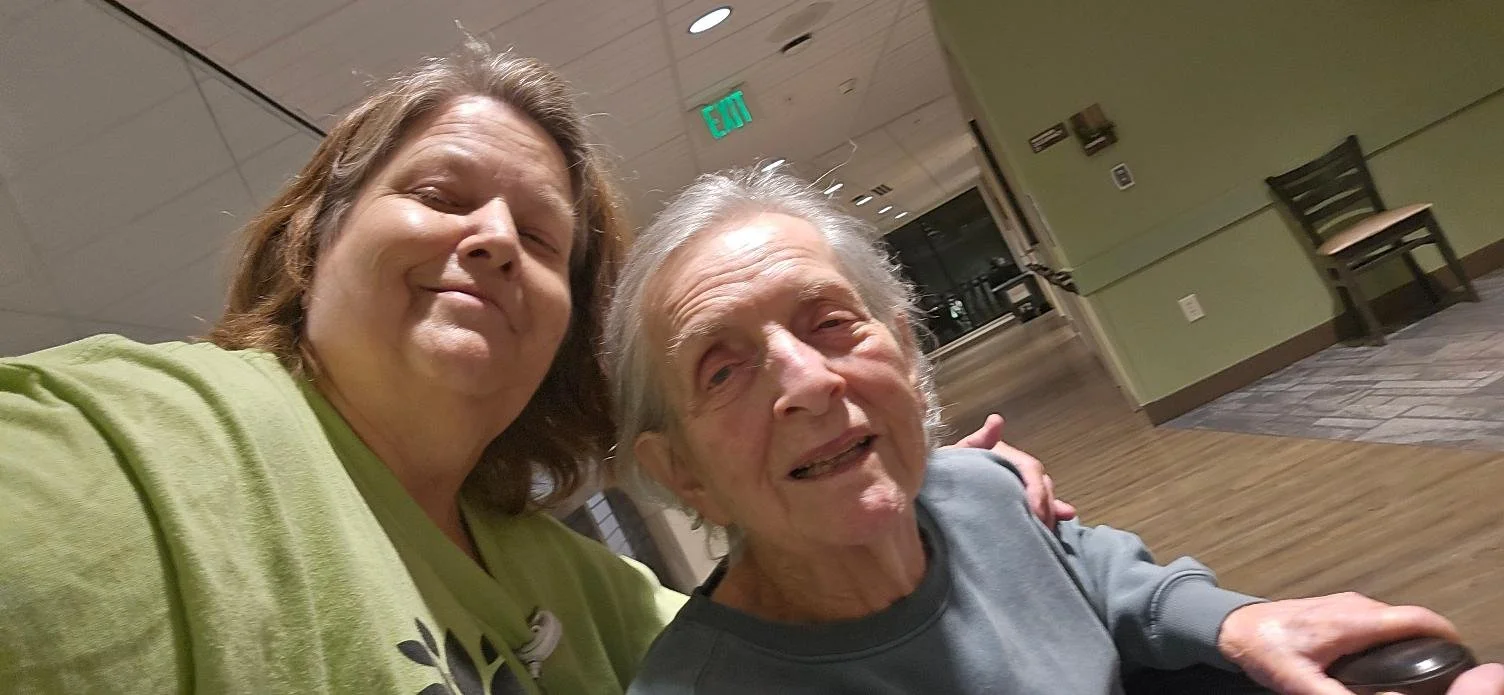 A woman and an elderly man smiling at the camera, taking a selfie together in an indoor setting with chairs and a hallway in the background.