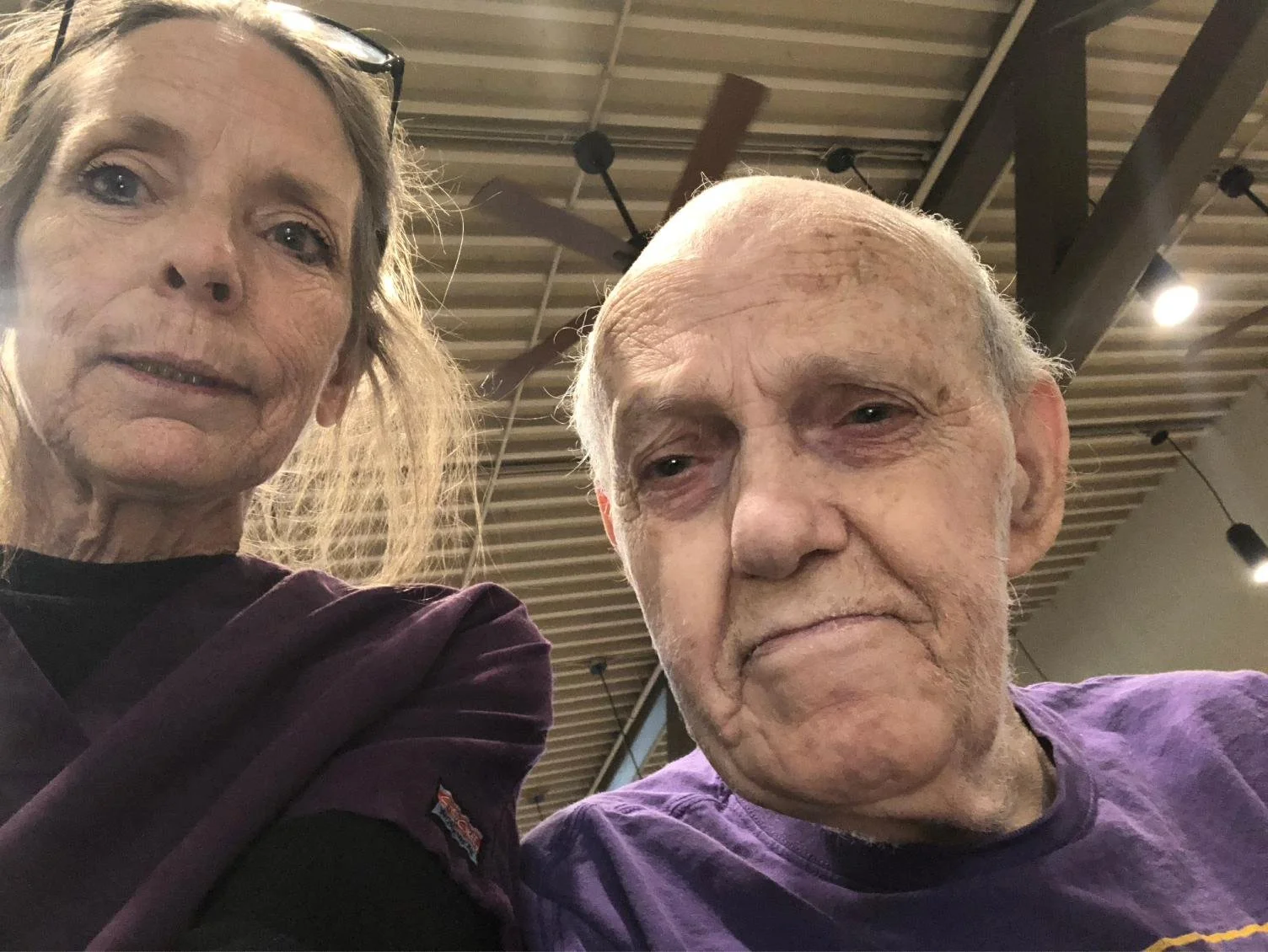 A selfie of an elderly woman and man in an indoor setting with wooden ceiling beams and hanging black lights. The woman has light skin, grey hair, and is wearing sunglasses on her head, looking directly at the camera with a neutral expression. The ma