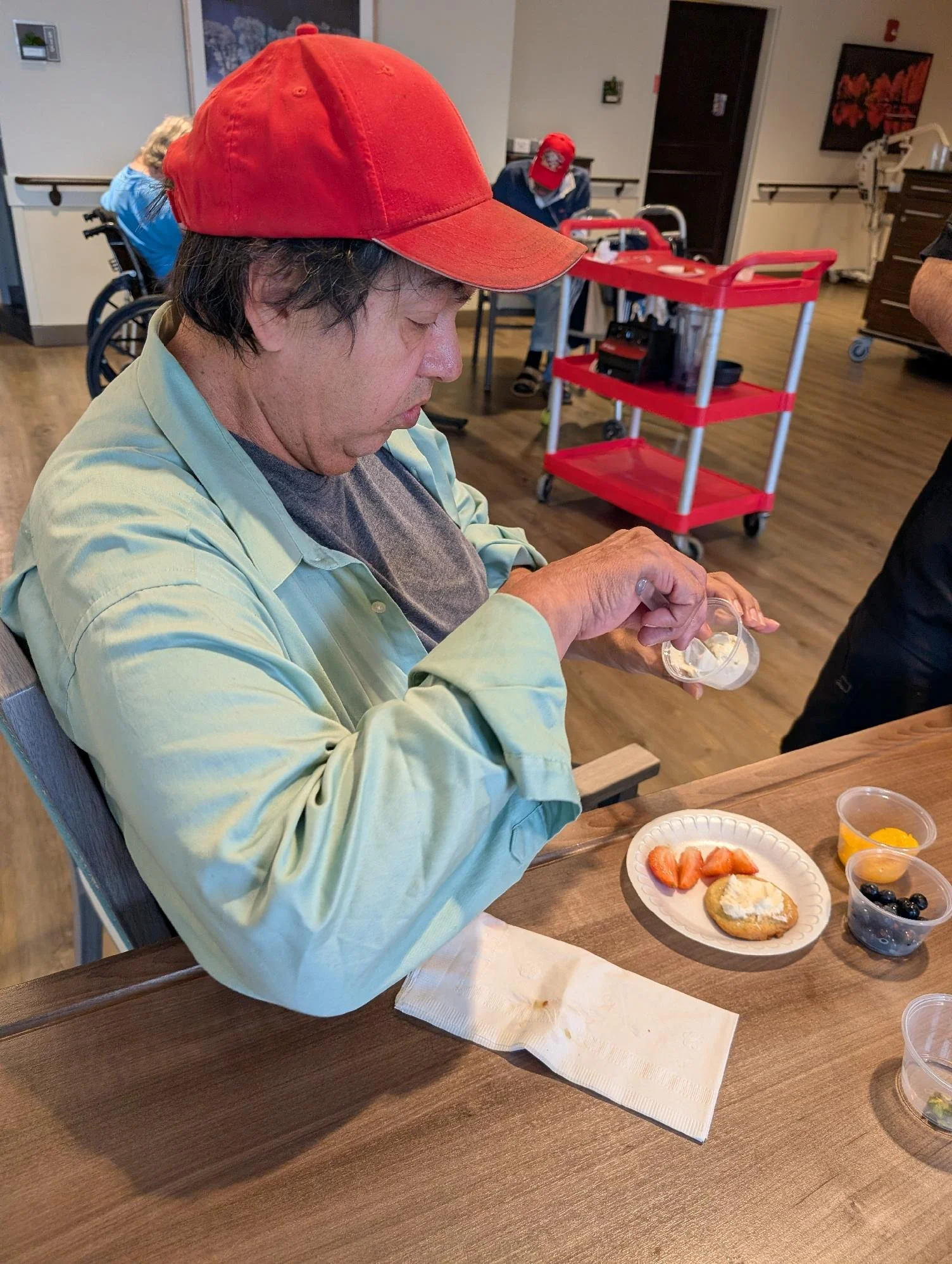 A man wearing a red cap and a light green shirt is sitting at a table with a plate of breakfast items, including strawberries, blueberries, a biscuit with cream cheese, and a small container of orange juice. In the background, other people are in a r
