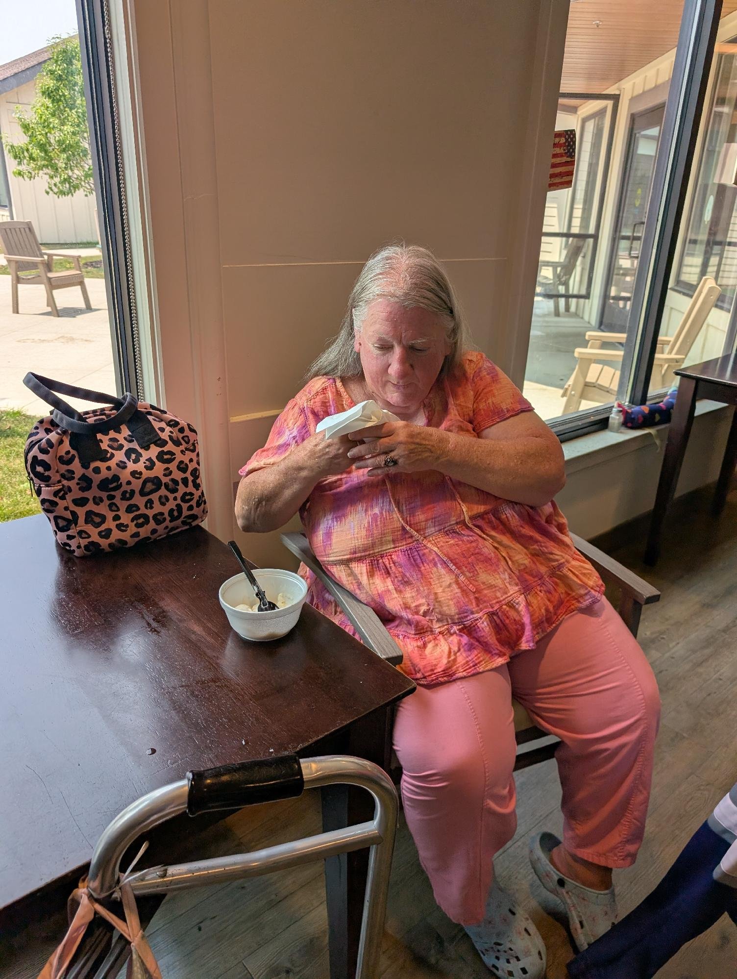 An elderly woman with long gray hair, wearing a colorful blouse and pink pants, is sitting in a chair near a window, looking at something in her hands. She has a patterned handbag on the table beside her and a walker is visible in the foreground.