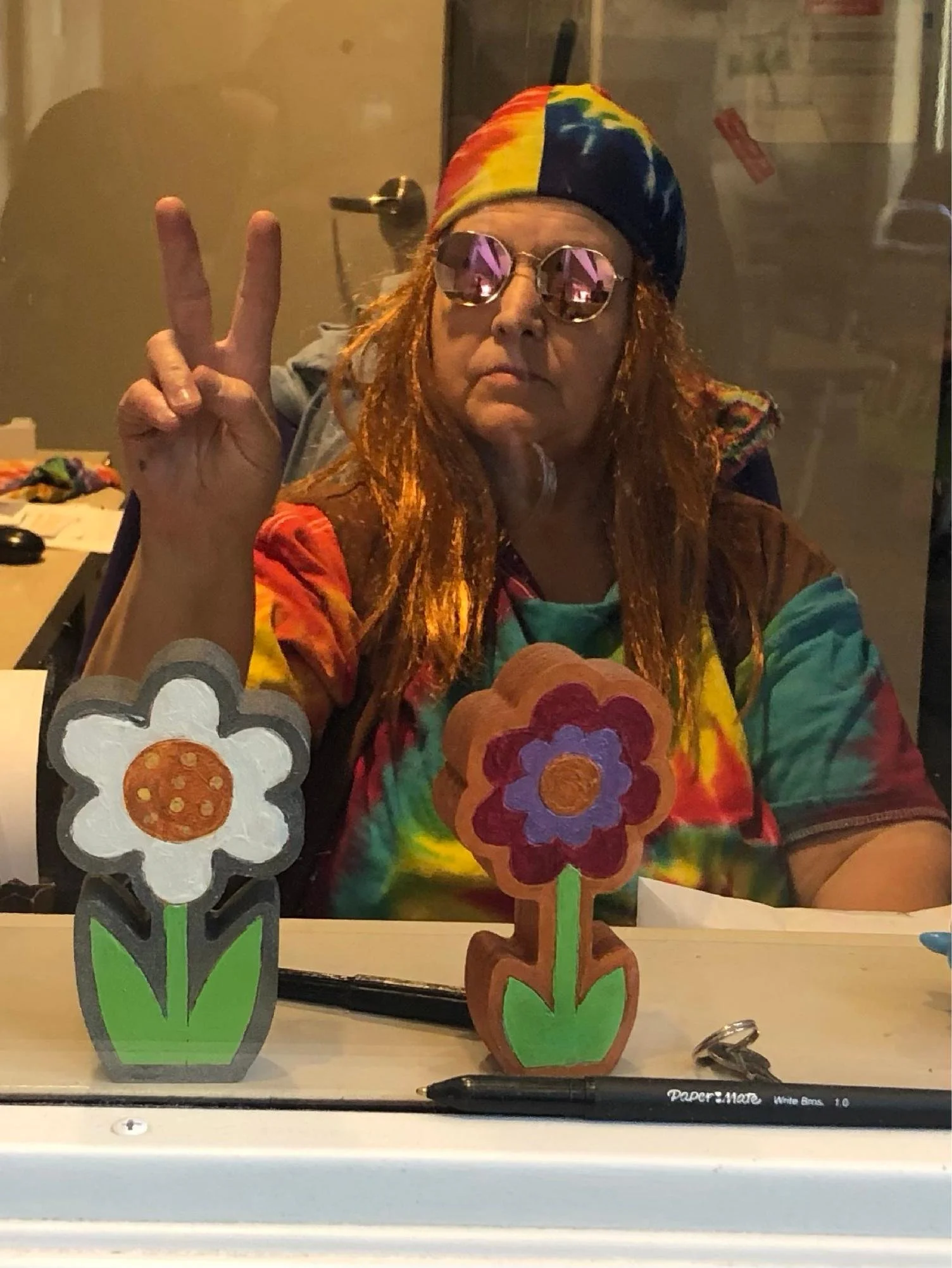 A person wearing a colorful tie-dye shirt, a rainbow-colored bandana, and round sunglasses making a peace sign with their hand. In front of them are three painted wooden flower decorations in pots.