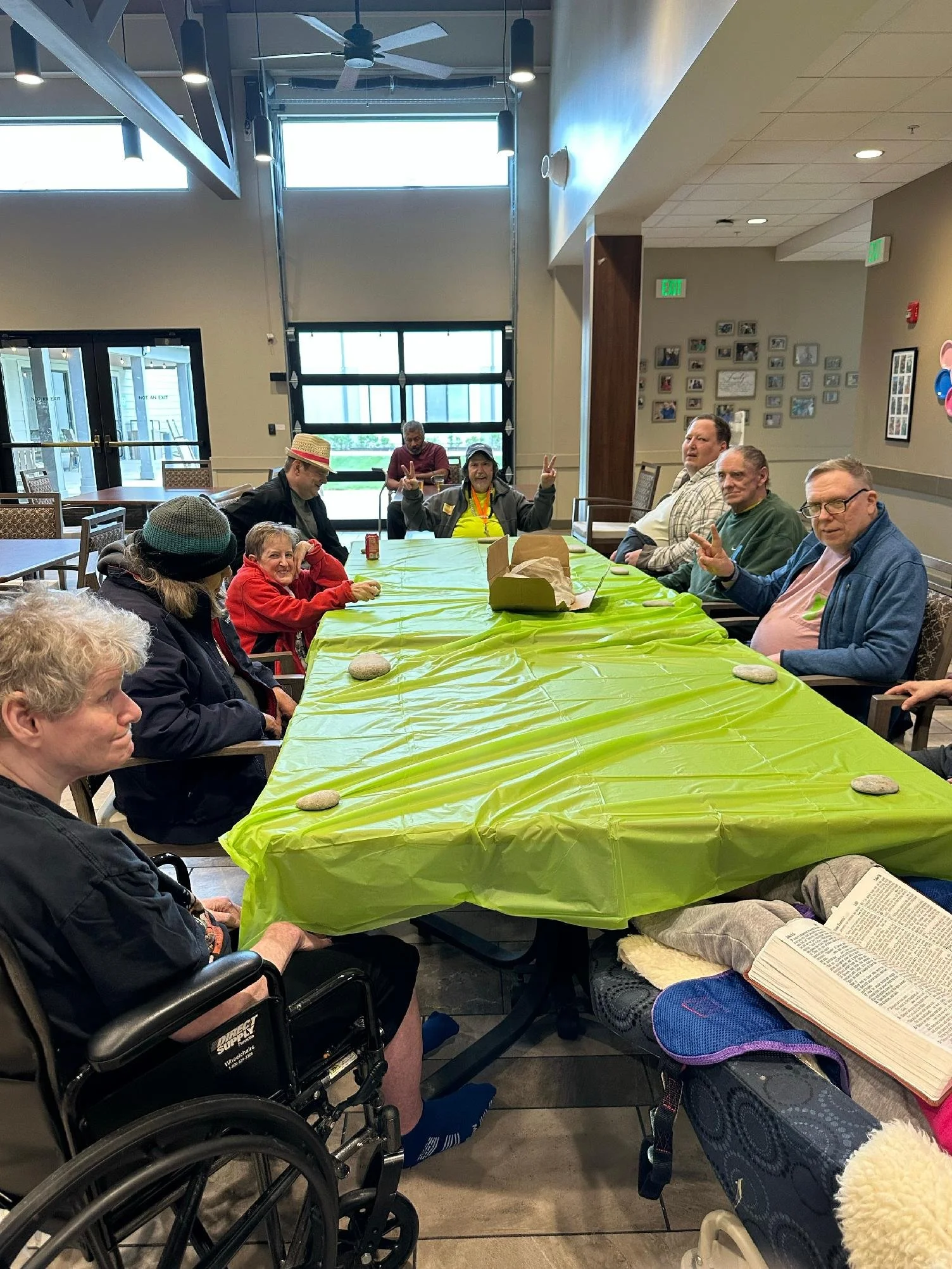 A group of people seated around a table covered with a bright green tablecloth, indoors in a well-lit room. Some are making peace signs, others are smiling or reading. One person in the foreground wears a wheelchair and a hat, and there is a woman wi
