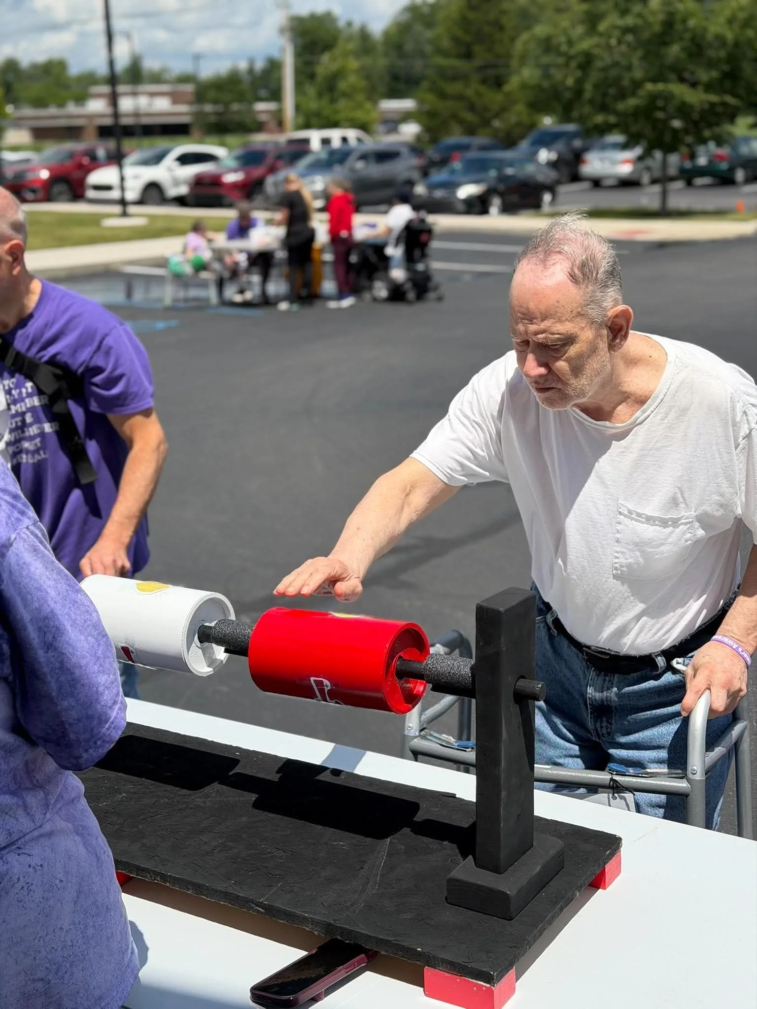 A man with a white shirt is using a domino effect machine, with a person in purple nearby, outdoors in a parking lot with cars and trees in the background, and several people gathered around a table in the distance.