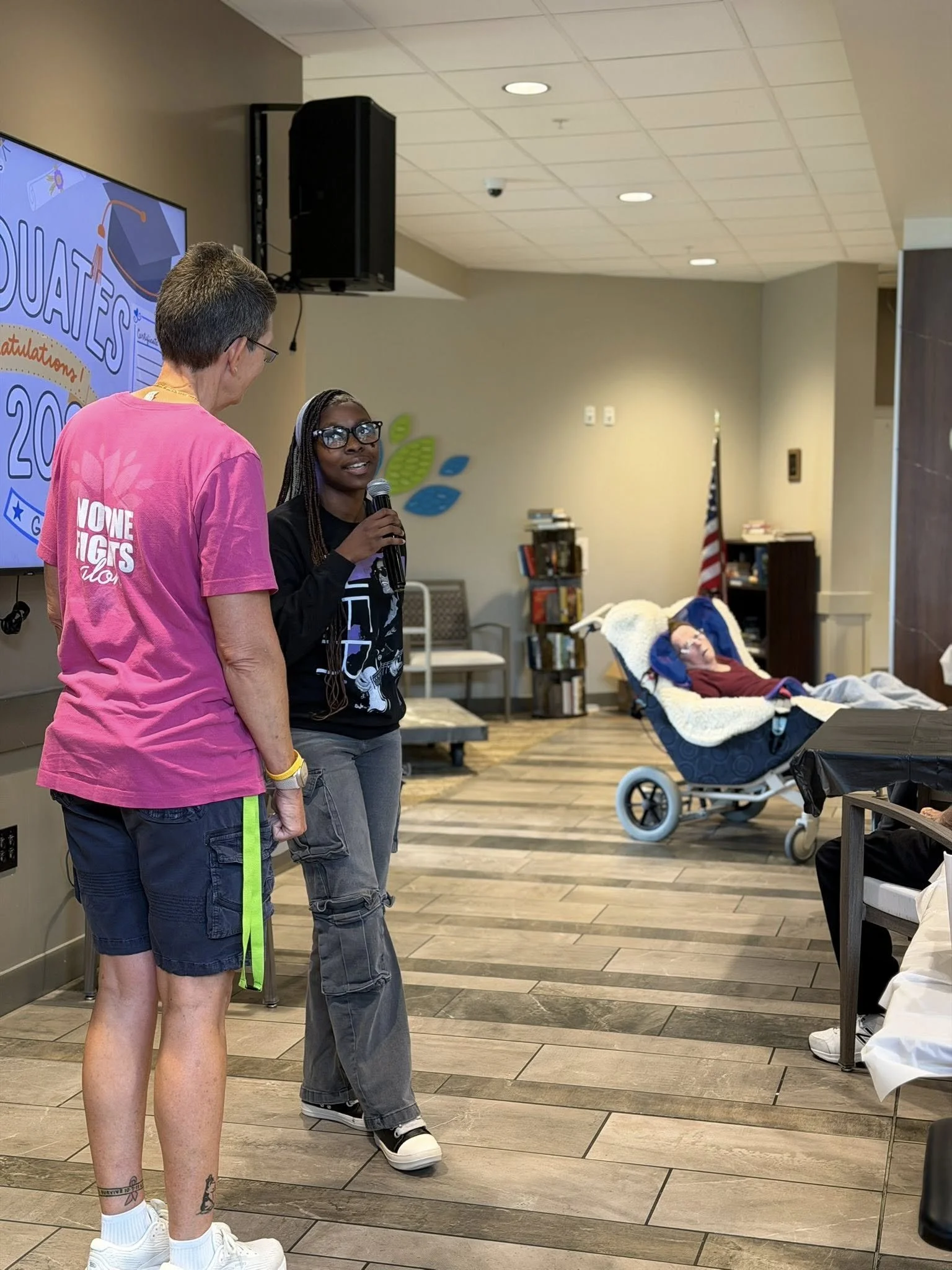 Two women stand and talk while one holds a microphone, inside a room with a large screen displaying a graduation-themed graphic. A person in a wheelchair is visible in the background.