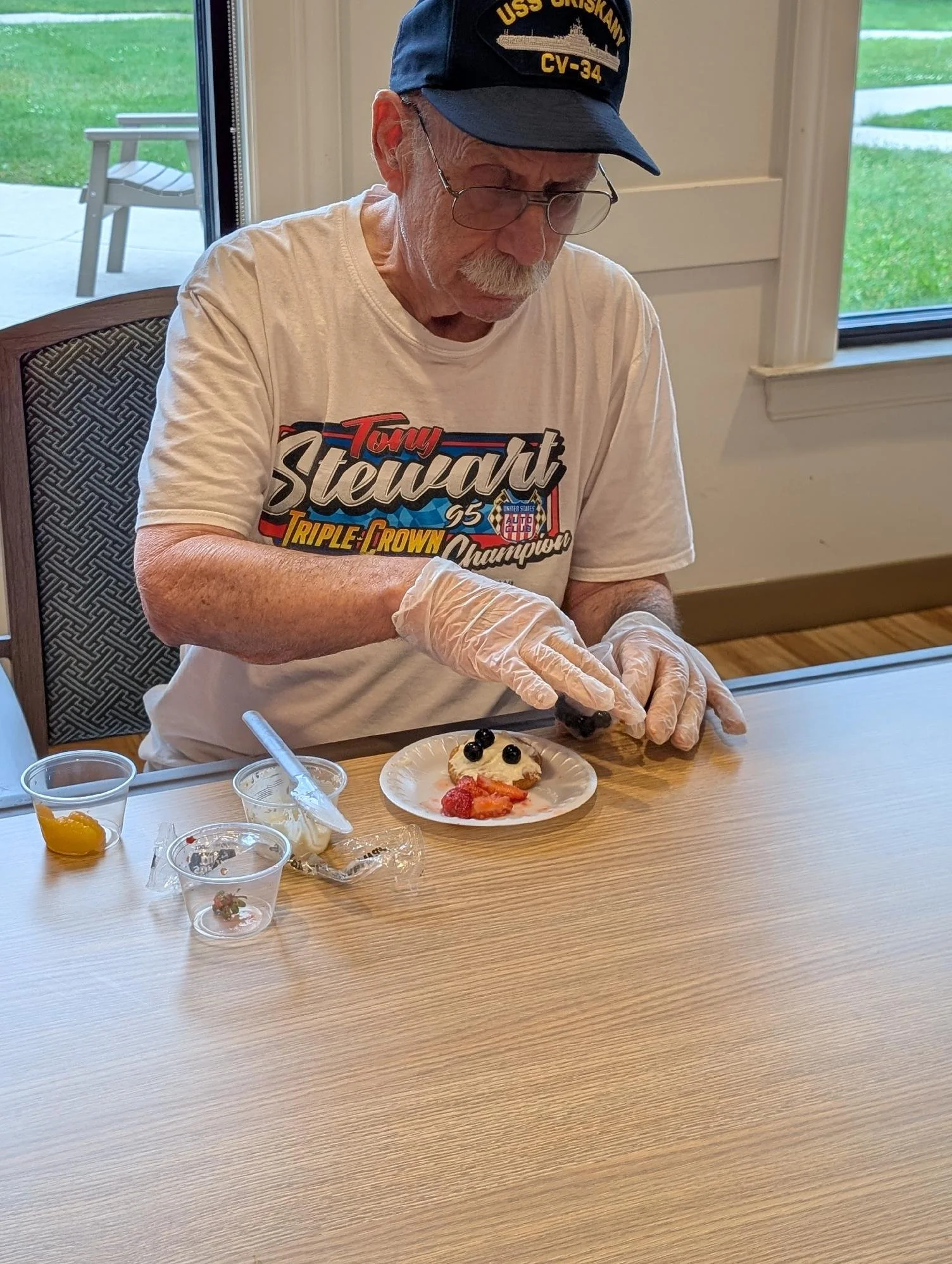 An elderly man with glasses and a navy hat labeled USS Griskkawk CV-34 is sitting at a table, preparing a dessert topped with blueberries. There are various small cups with toppings or ingredients on the table, and a window in the background shows a 