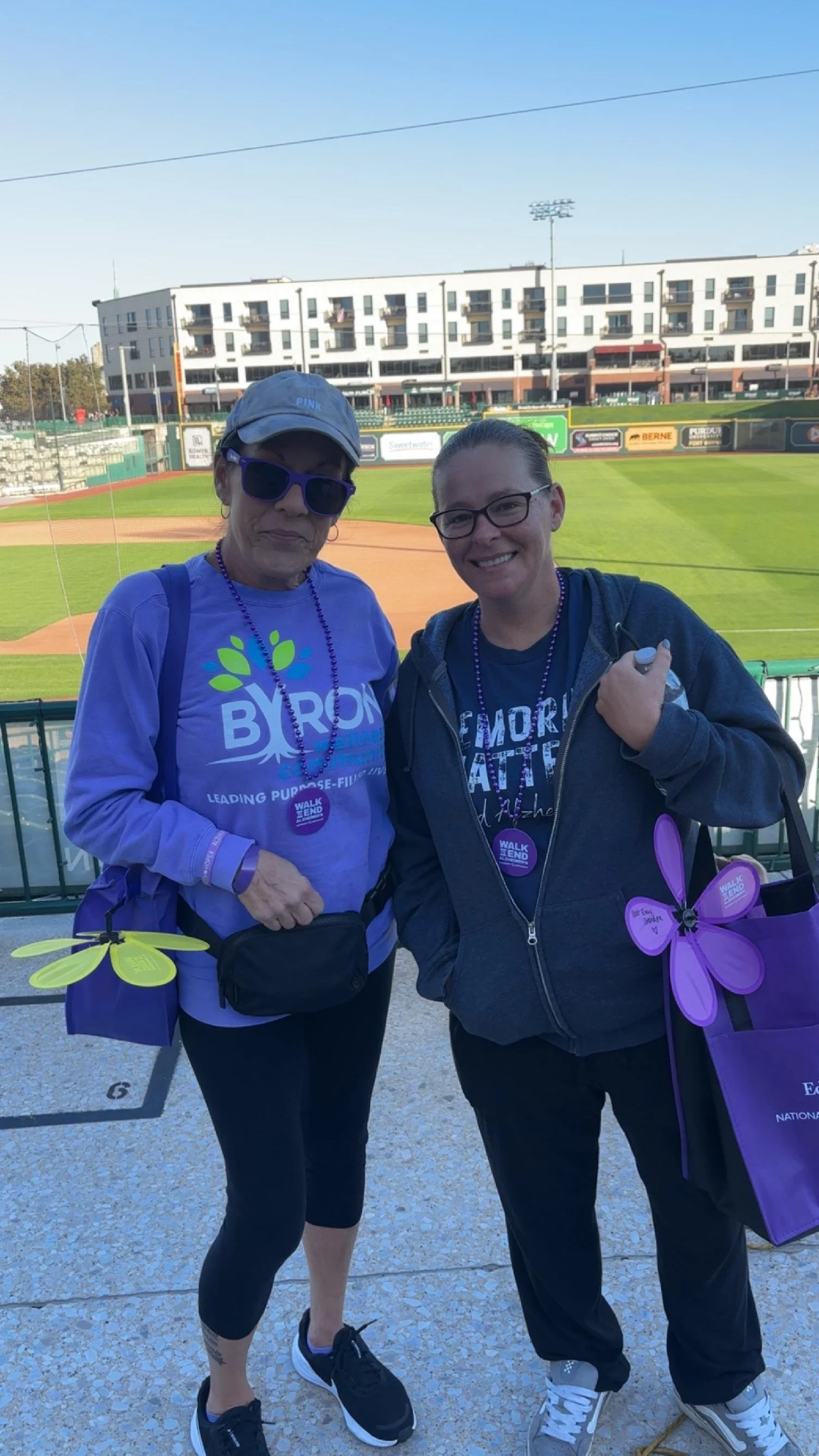 Two women standing in front of a baseball field, smiling at the camera, wearing purple shirts with purple beaded necklaces, and holding purple and yellow pinwheels, with stadium seats and a white residential building in the background.