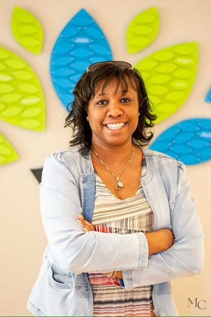 Smiling woman with crossed arms standing in front of colorful leaf-shaped wall decorations.