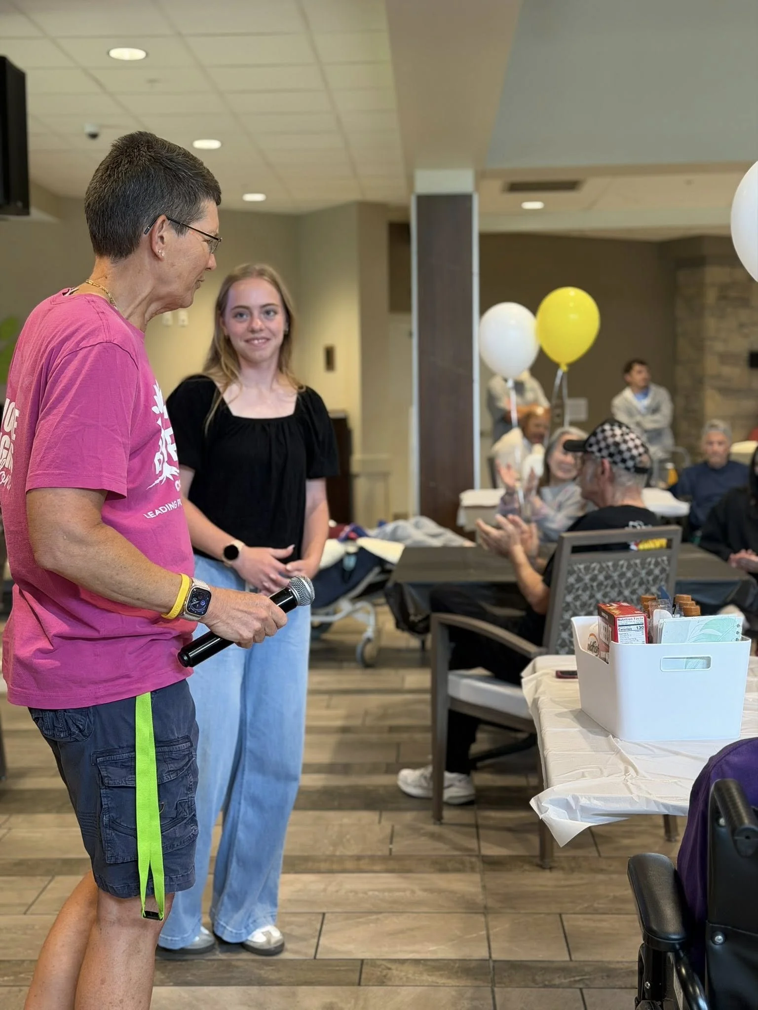 A man wearing sunglasses, a pink t-shirt, and navy shorts holding a microphone standing next to a young woman in black top, in a room with people sitting and standing, some holding balloons, and tables with food and supplies.