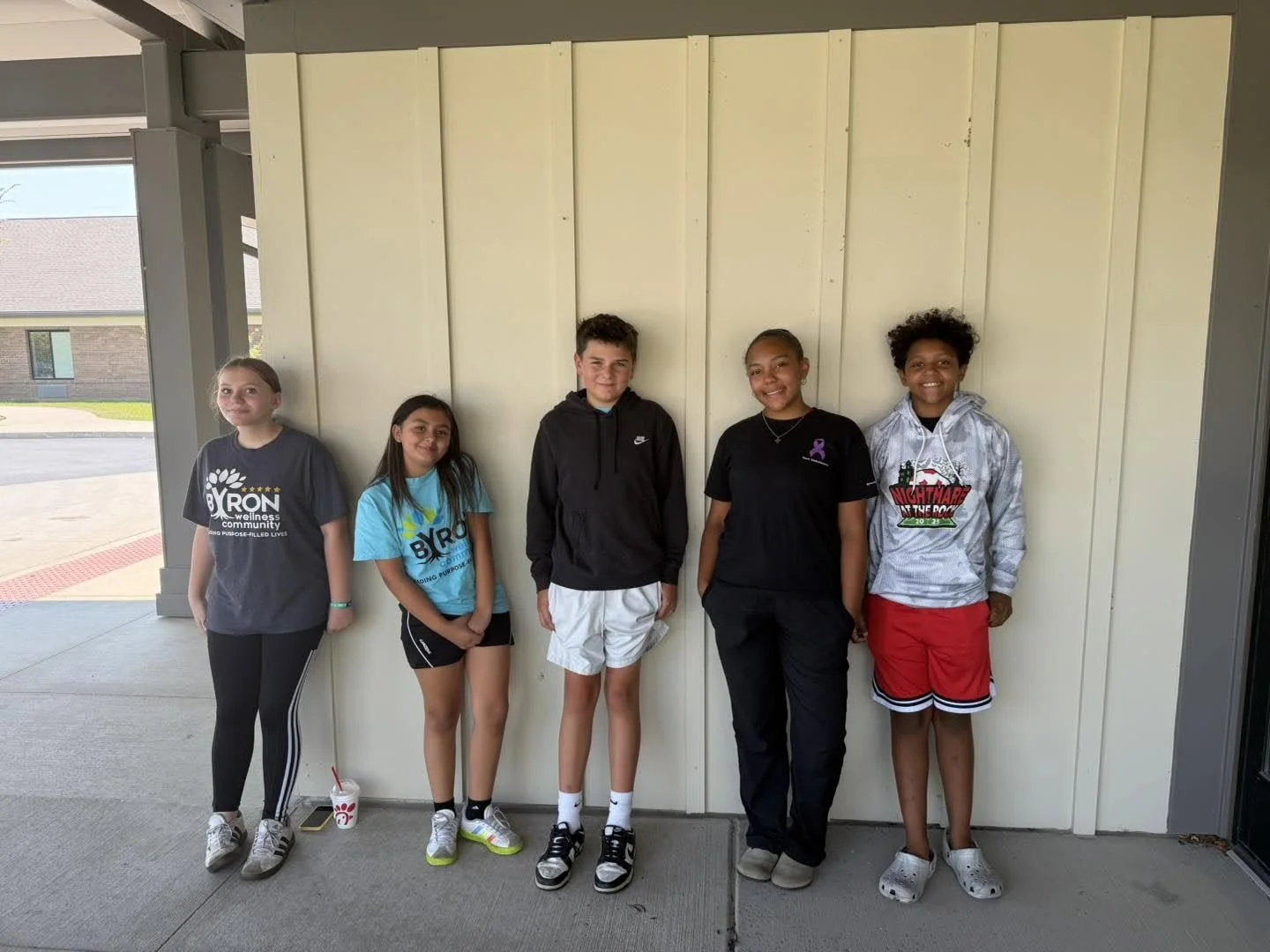 Five children standing in a row outdoors against a beige wall, smiling, dressed in casual athletic clothing.