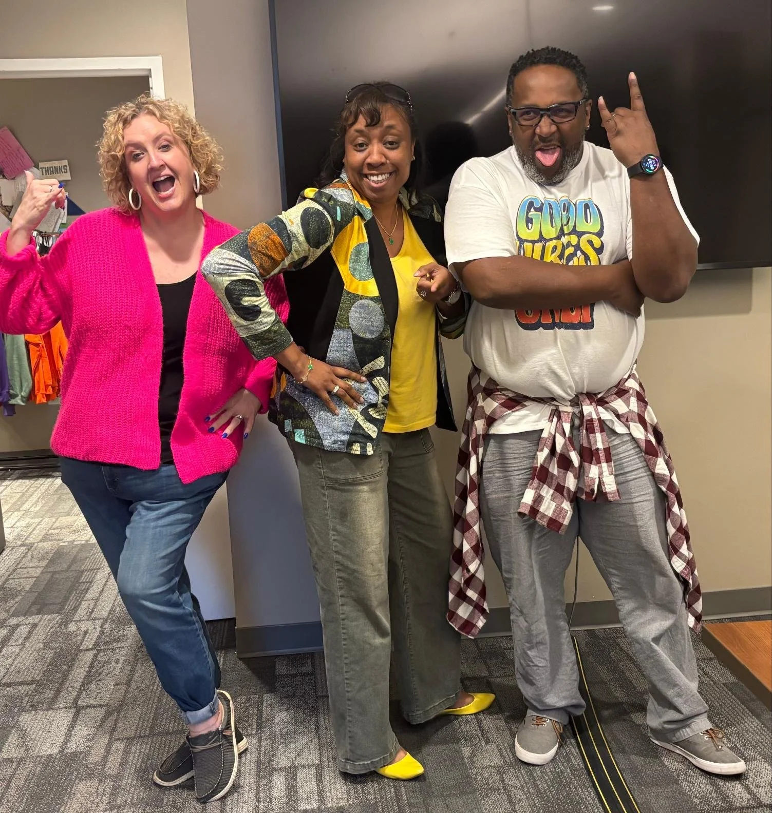 Three people standing together in a room, smiling and making playful gestures. The woman on the left is wearing a bright pink sweater and comfortable jeans. The woman in the middle is wearing a patterned blazer and yellow top. The man on the right is