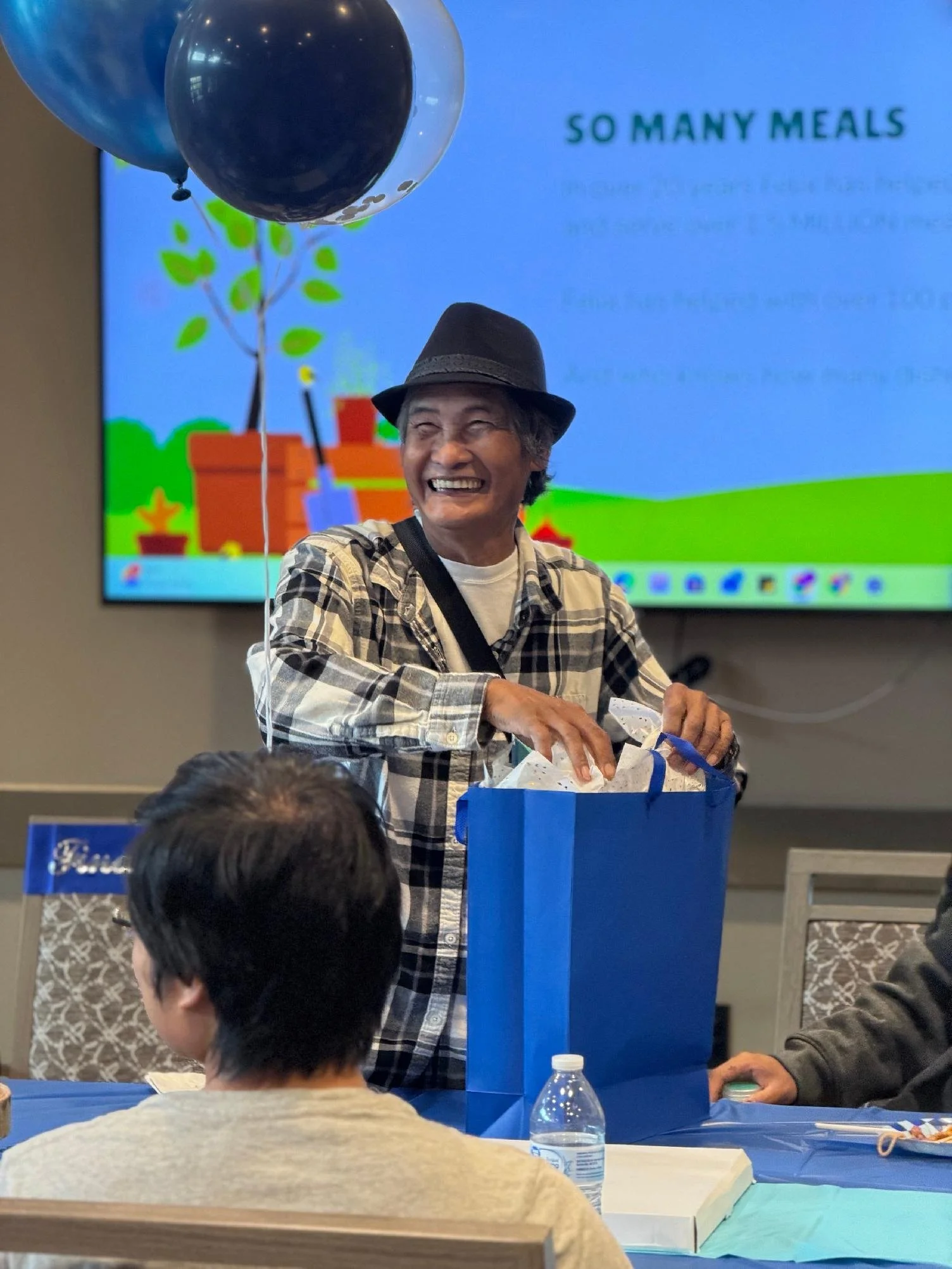 A smiling elderly woman wearing a hat and plaid shirt is standing at a table, with balloons and a large blue gift bag in front of her, during a celebration or event.