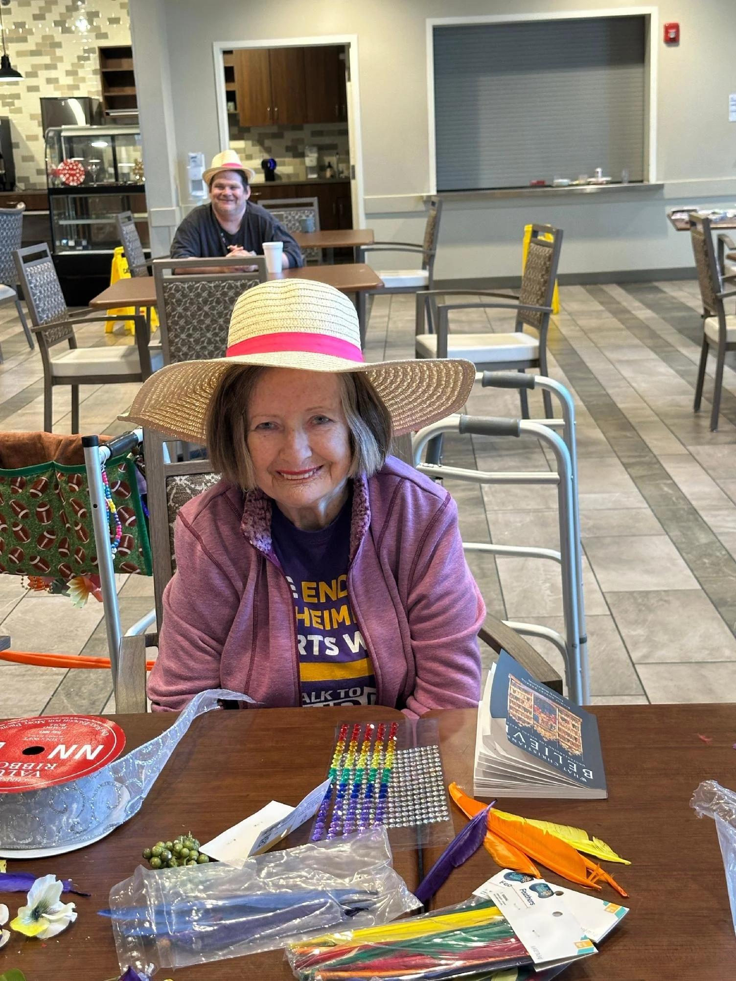 A smiling elderly woman wearing a large straw hat with a pink band, sitting at a table in a cafe or community center. The table has craft supplies such as colorful feathers, beads, and stickers. Behind her, a man wearing a straw hat is sitting at ano