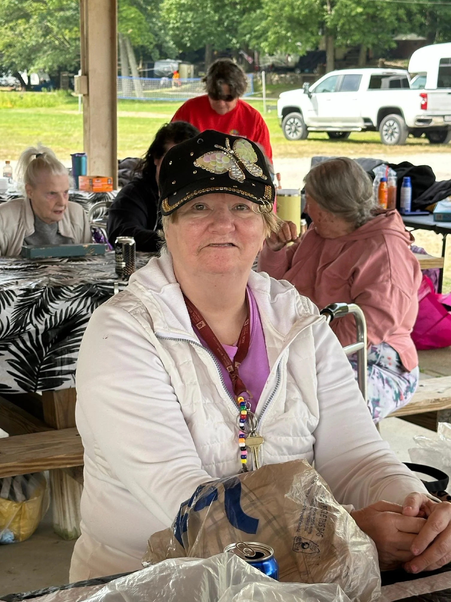 A group of people gathered outdoors at a picnic, sitting at picnic tables under a pavilion with trees and parked cars in the background. The focus is on a smiling woman wearing a black cap with a butterfly design and colorful beads.