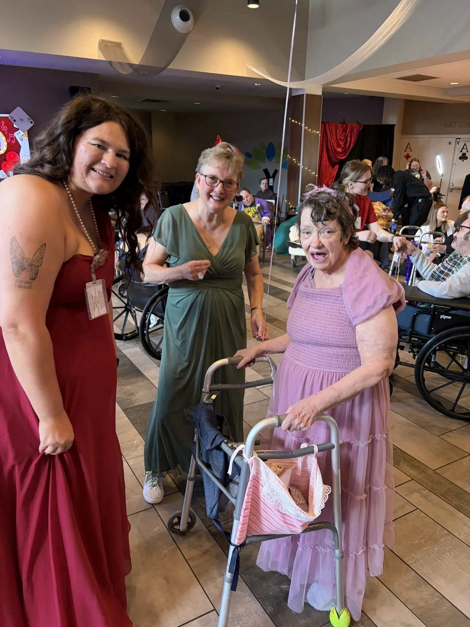 Three women smiling and holding hands in a celebration, with other elderly people seated at tables in the background. One woman is using a walker, and there are birthday decorations and a red curtain in a lively indoor setting.