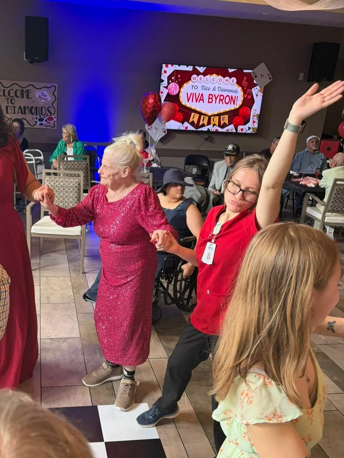 Older woman dancing with a younger woman at a celebration, others seated around, large digital screen in the background displaying 'Welcome to Dice & Diamonds Viva Byron!'