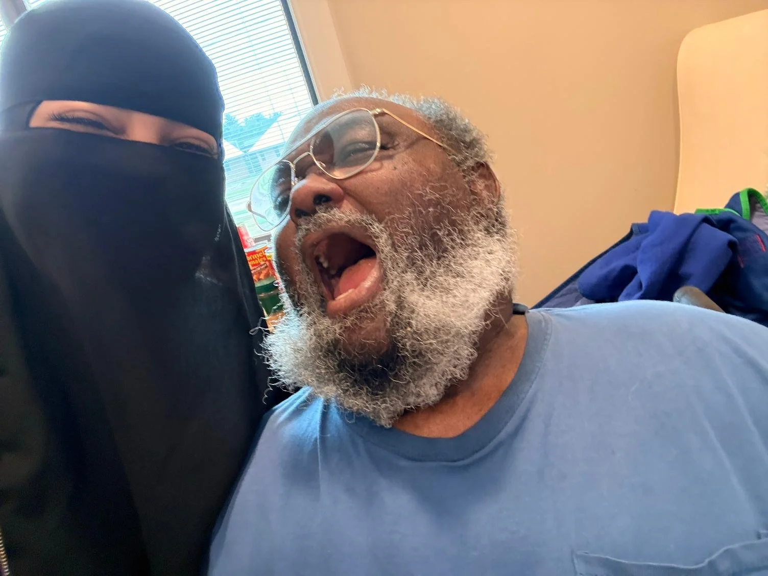 A man with glasses and a gray beard leaning his head against a woman wearing a black face mask and head covering, both smiling and appearing joyful in an indoor setting.