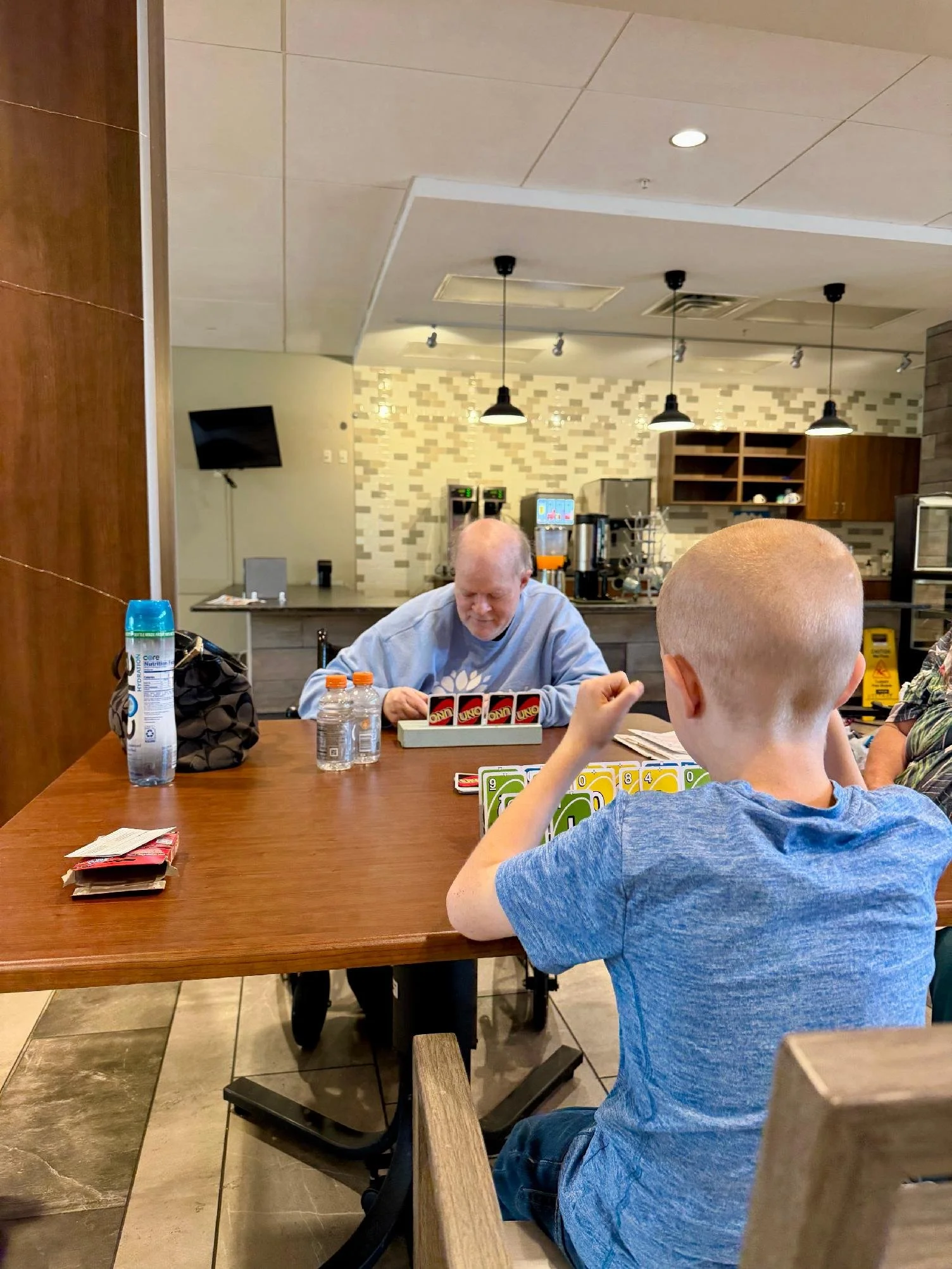 Two boys sitting at a wooden table playing Uno card game in a restaurant. The boy in the foreground has a shaved head and is wearing a blue shirt. The older boy seated across has a bald head and is wearing a light blue hoodie. There are water bottles