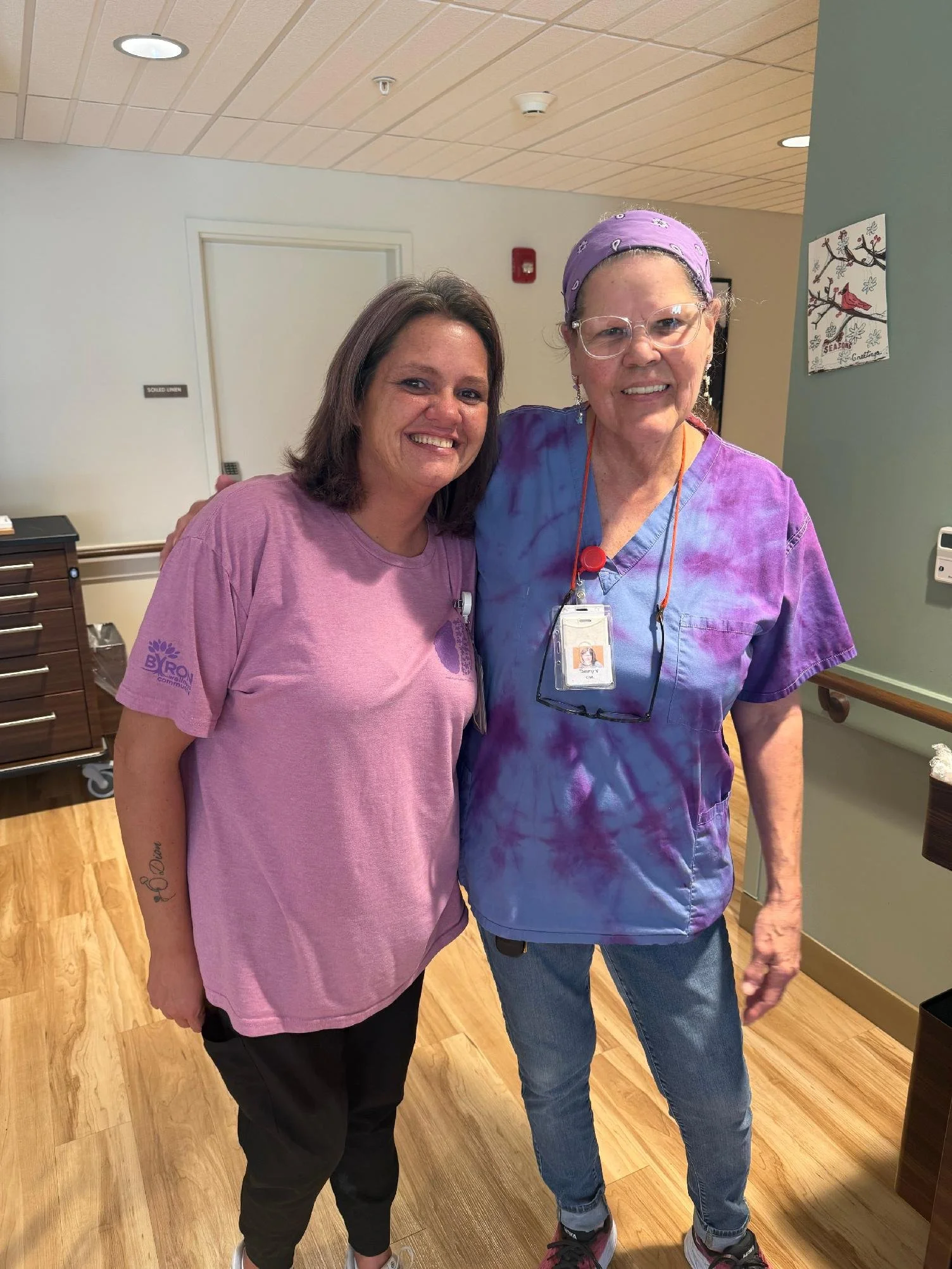 Two women standing close together indoors, smiling at the camera. One woman has shoulder-length brown hair and is wearing a pink T-shirt and black pants. The other woman has short gray hair, glasses, a purple bandana, and is dressed in purple scrubs 
