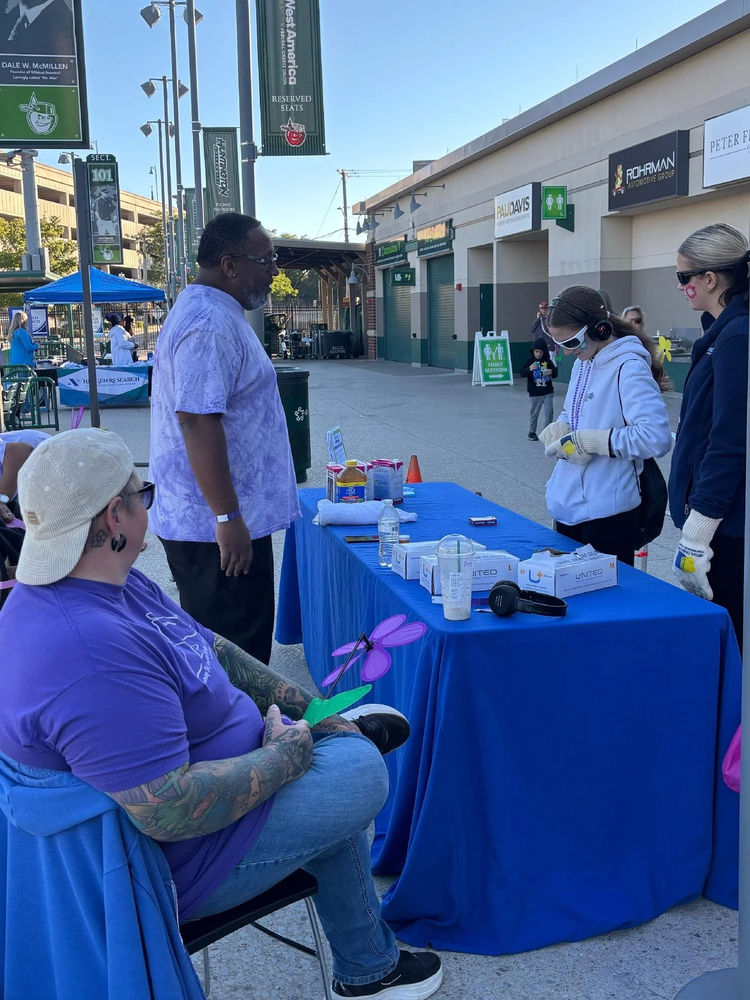 A group of people at an outdoor event with a table covered in a blue cloth, medical supplies like face masks, hand sanitizer, and gloves, and a person sitting with arms crossed and wearing a purple T-shirt and glasses. There are others standing and e