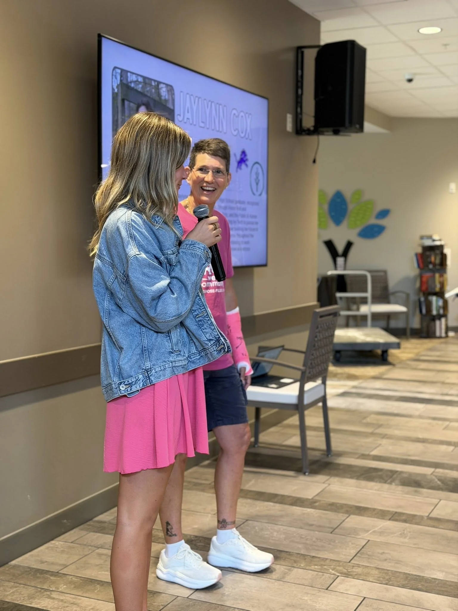 Two women standing indoors, one with blonde hair wearing a denim jacket and pink dress holding a microphone, and the other woman with short hair in a pink shirt and black shorts smiling. There is a large screen behind them with text, a wall decoratio