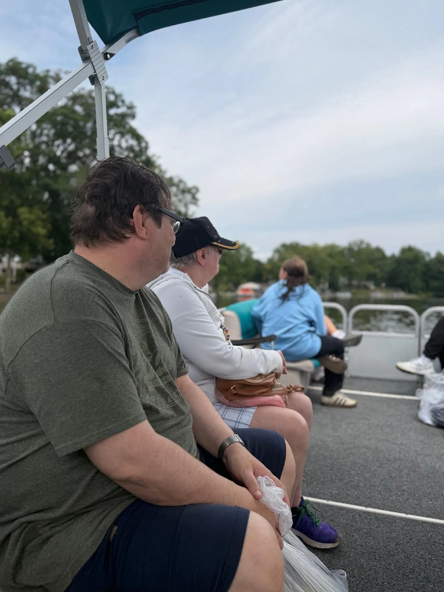 People sitting on benches outdoors by a body of water, with trees and cloudy sky in the background.