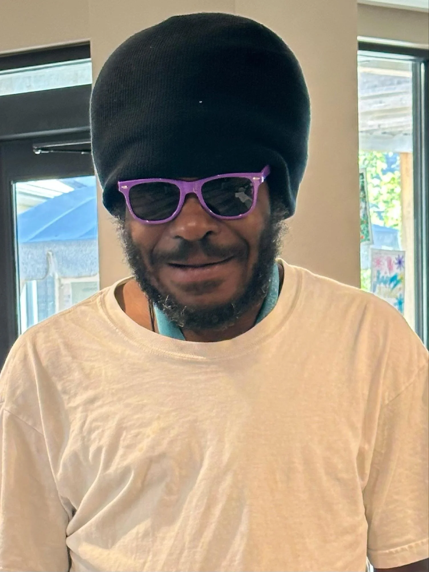 A man smiling, wearing a black beanie and purple sunglasses, dressed in a light-colored T-shirt, indoors near sunny windows.