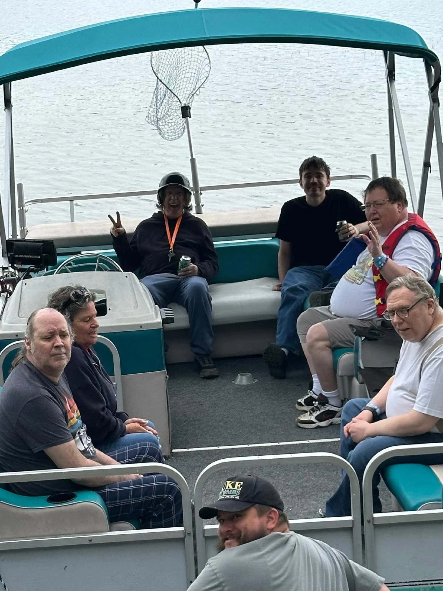 Group of people sitting and standing on a boat with water in the background, some smiling and holding drinks.