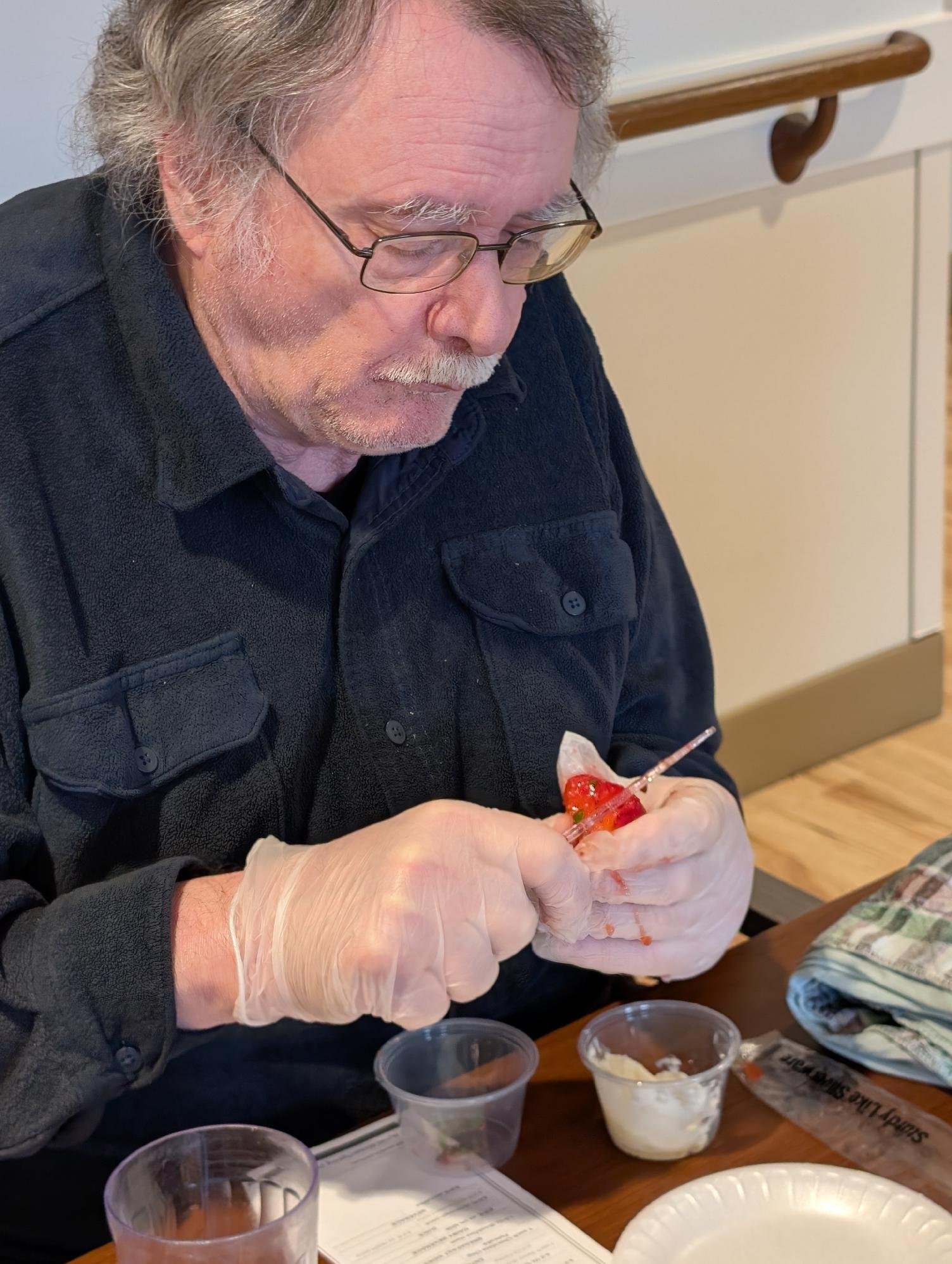 Older man with gray hair and glasses wearing black shirt and gloves preparing food, possibly strawberries, at a dining table with cups of ice cream, a paper plate, and a menu.