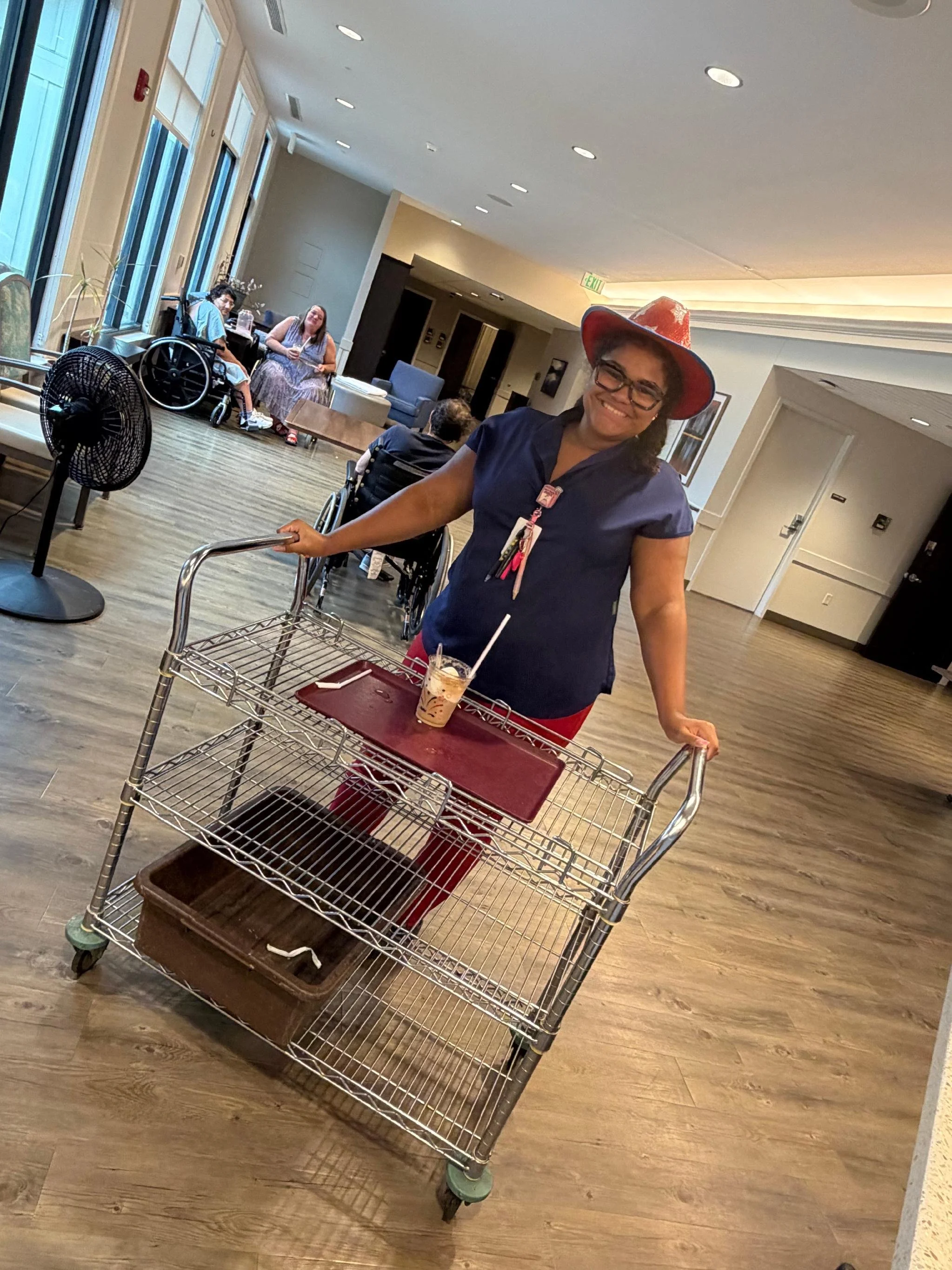 Smiling woman with glasses wearing a red cowboy hat pushing a shopping cart in a bright, modern indoor space. In the background, a person in a wheelchair and another woman sitting at a table are visible.