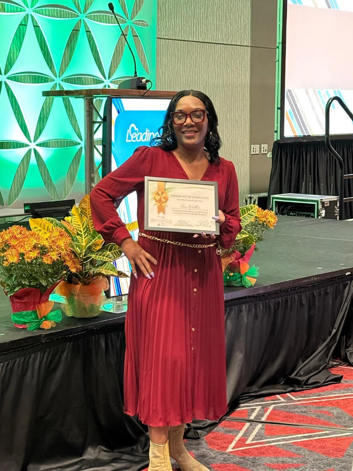 A woman in a red dress and cowboy boots holding a certificate, standing in front of a decorated stage with flowers, a screen, and a podium at an awards ceremony or event.