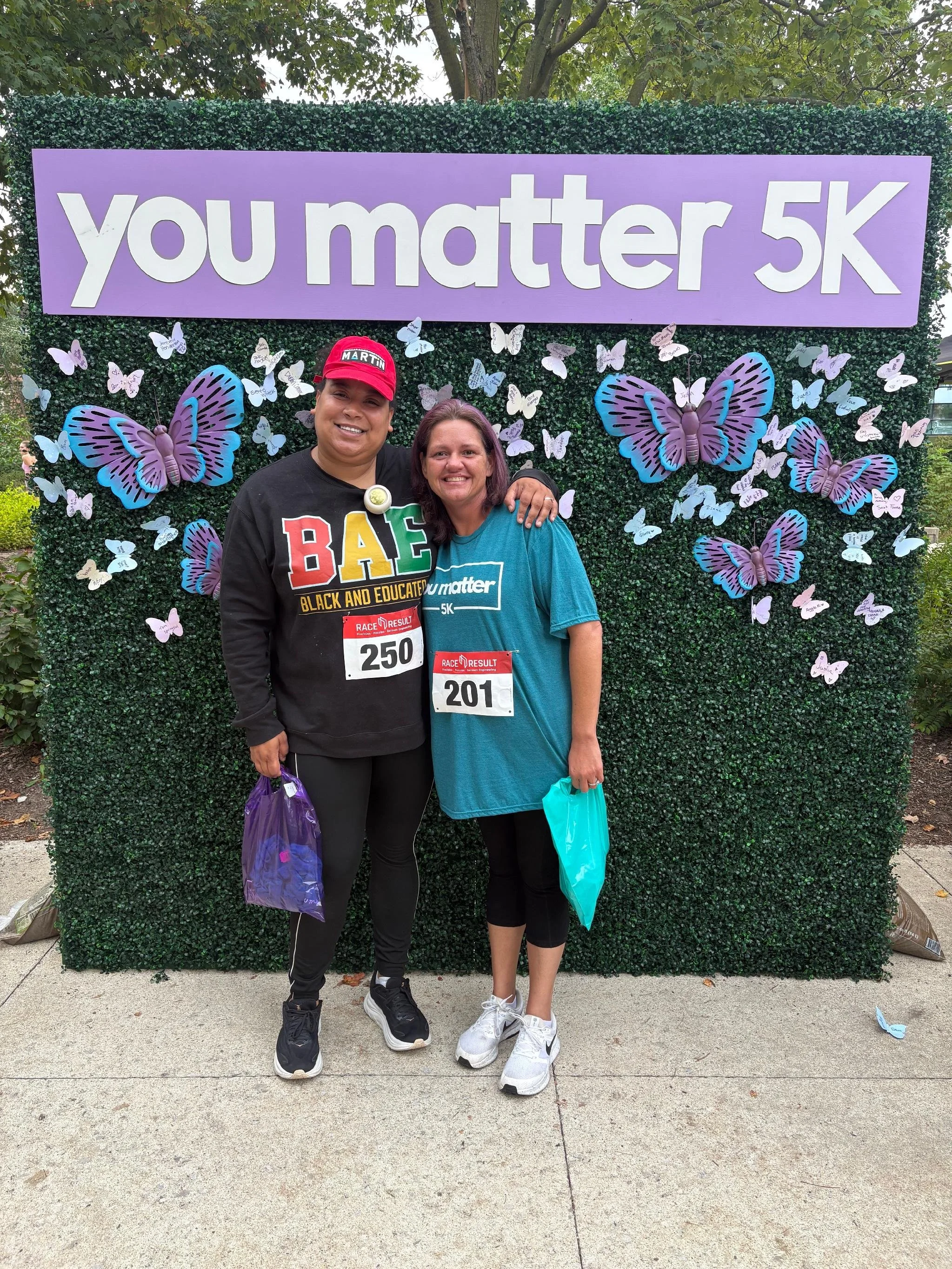 Two women standing in front of a green hedge with purple butterflies and 'you matter 5K' sign, holding purple and turquoise bags, wearing race bibs, smiling.