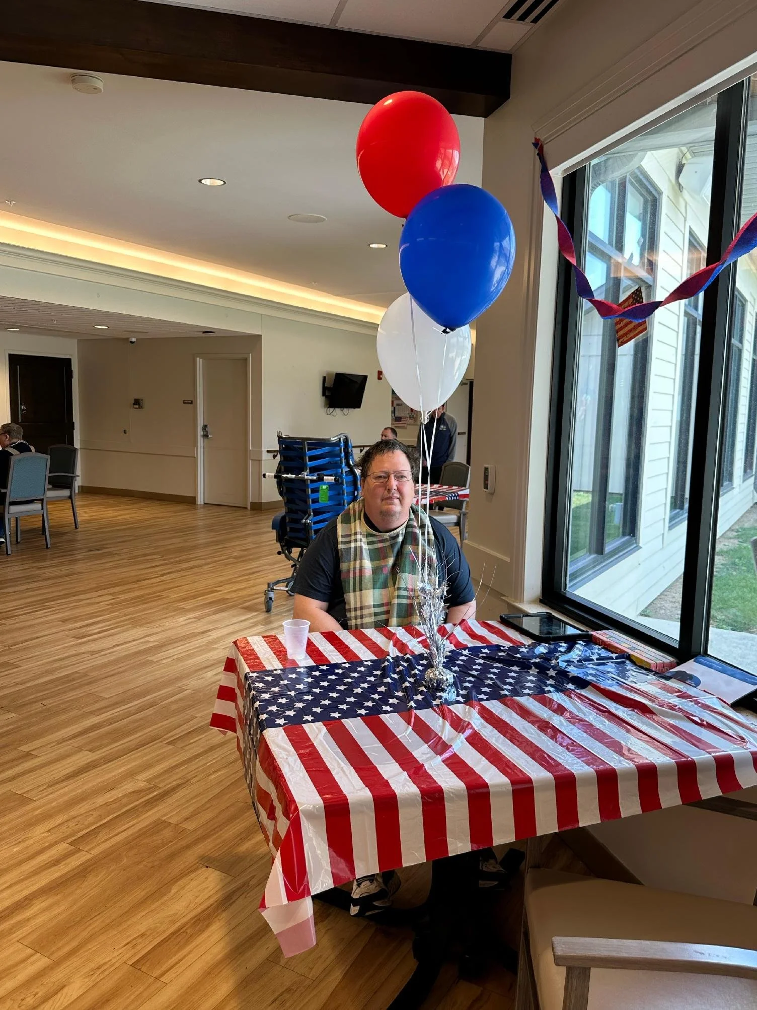 A man sitting at a table with a patriotic American flag tablecloth, with red, white, and blue balloons and decorations, in a decorated indoor setting with large windows showing a house outside.
