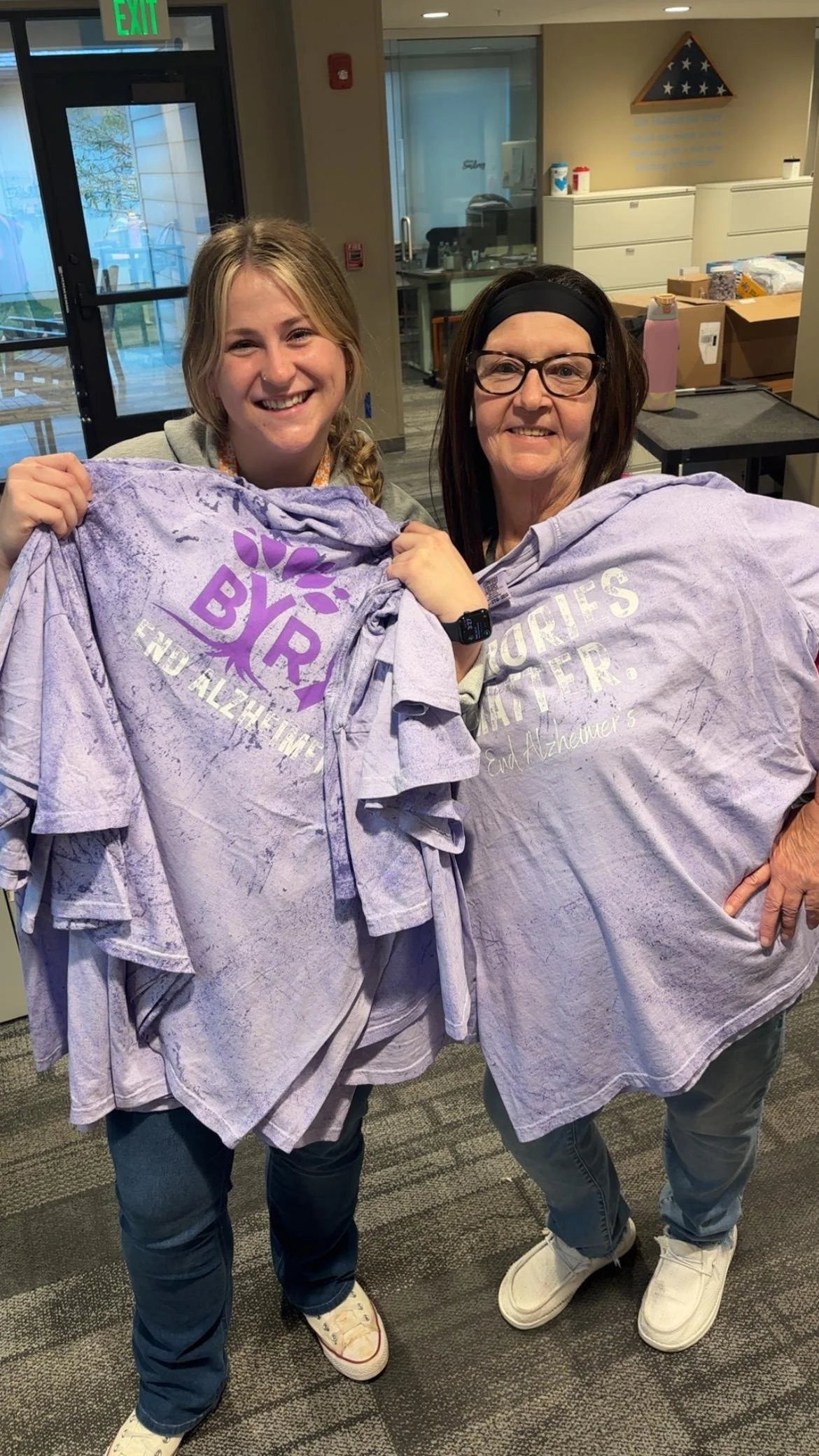 Two women standing indoors, holding up purple t-shirts with the text 'END ALZHEIMER'S' printed on them. They are smiling at the camera.
