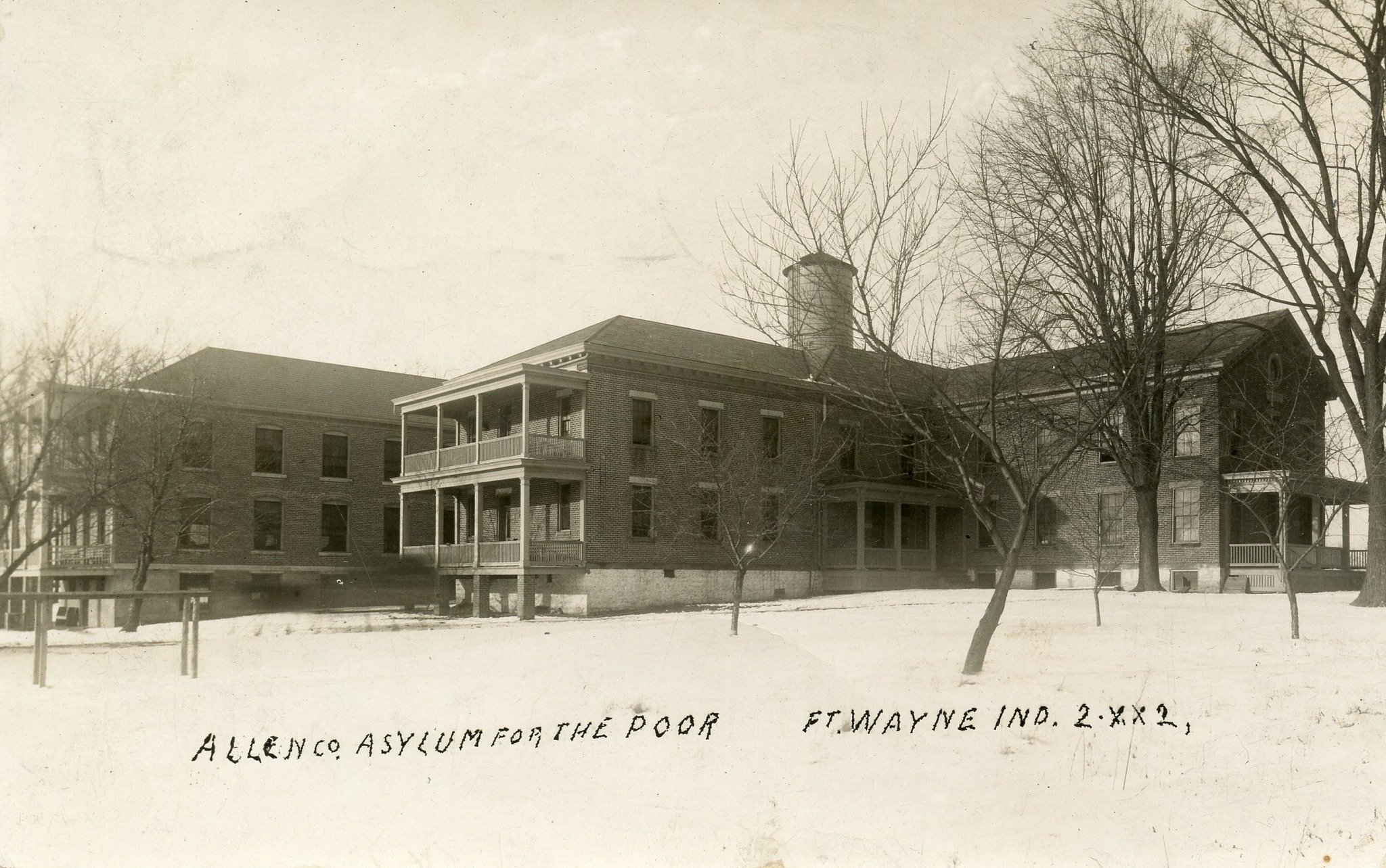 Black and white photo of a large brick building with a porch and trees in the foreground. The text on the photo reads: "ALLENCO ASYLUM FOR THE POOR FT. WAYNE IND. 2 X 2."