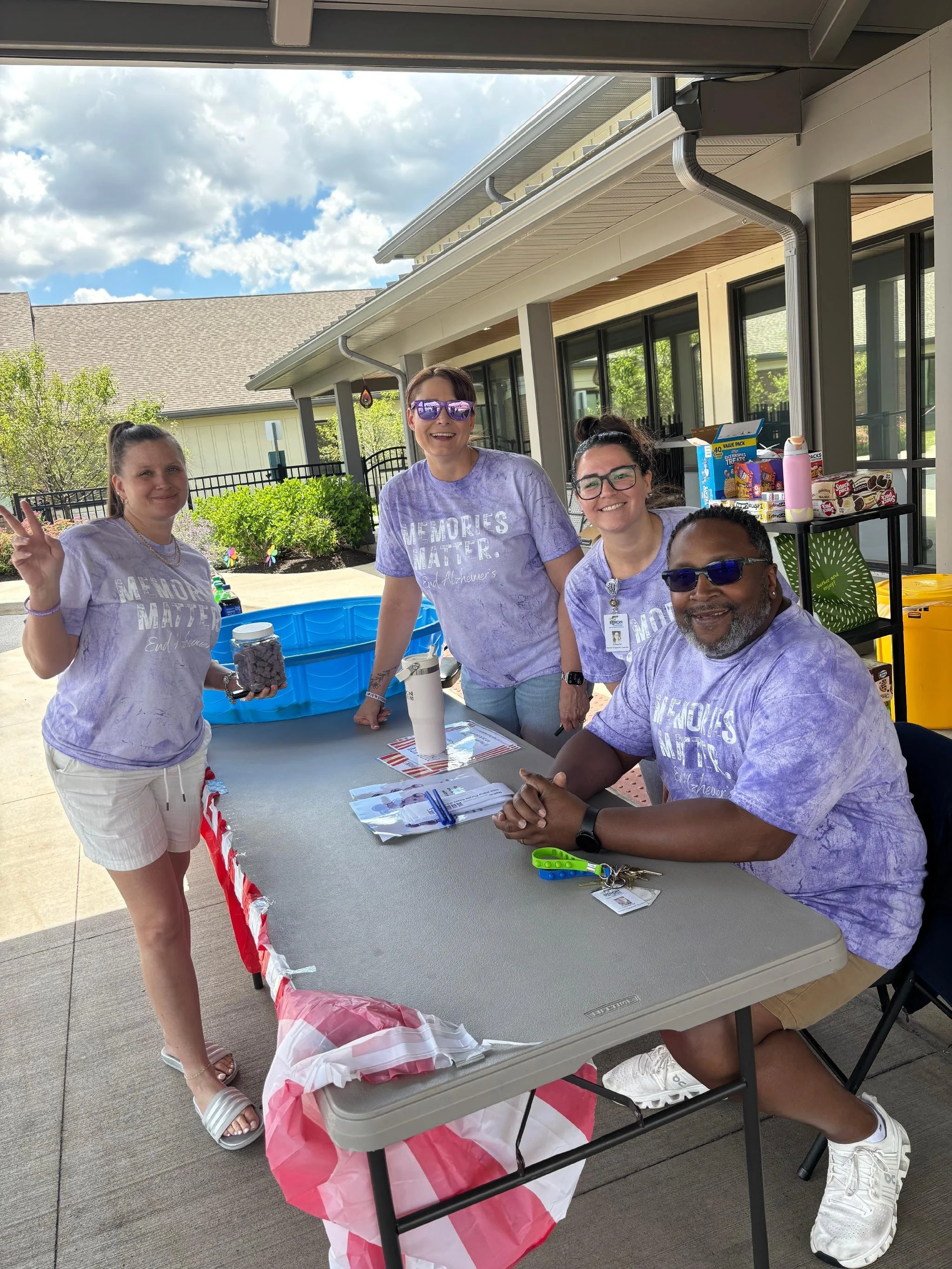 Group of four people standing and sitting around a table outdoors, smiling for the photo, with snacks and drinks on a table behind them. They are wearing purple shirts with white text that reads 'Memories Matter.' The setting appears to be a backyard