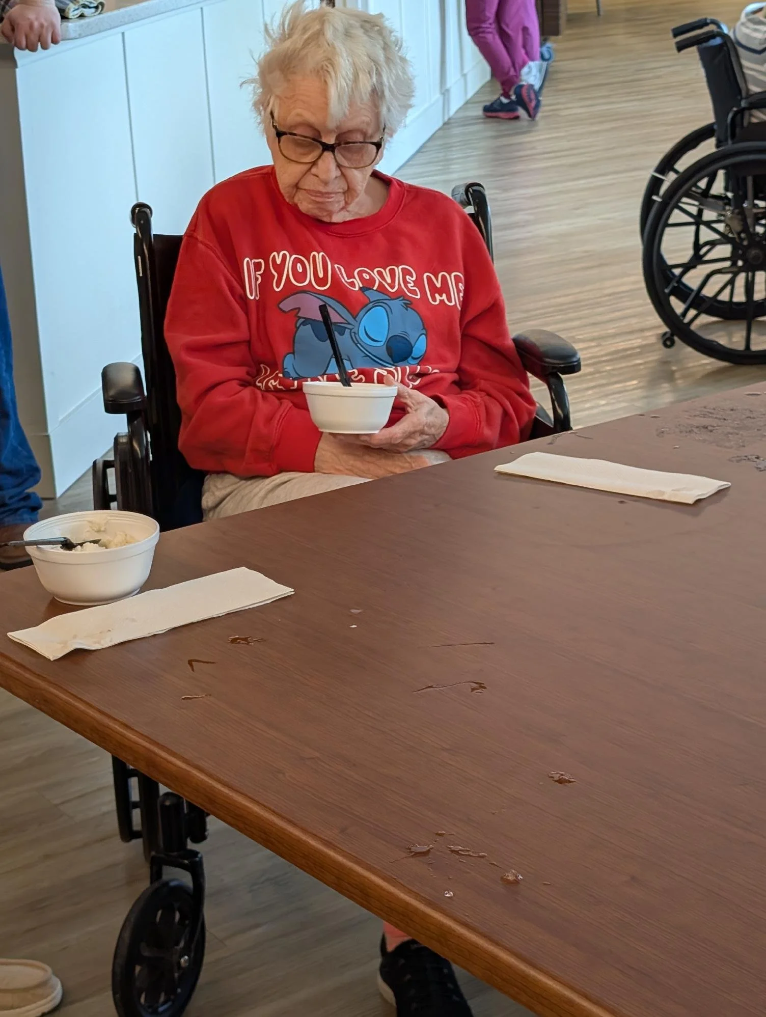 An elderly woman with white hair and glasses sitting in a wheelchair at a table, holding a bowl, with a bowl of food and napkin on the table in front of her in a casual dining area.
